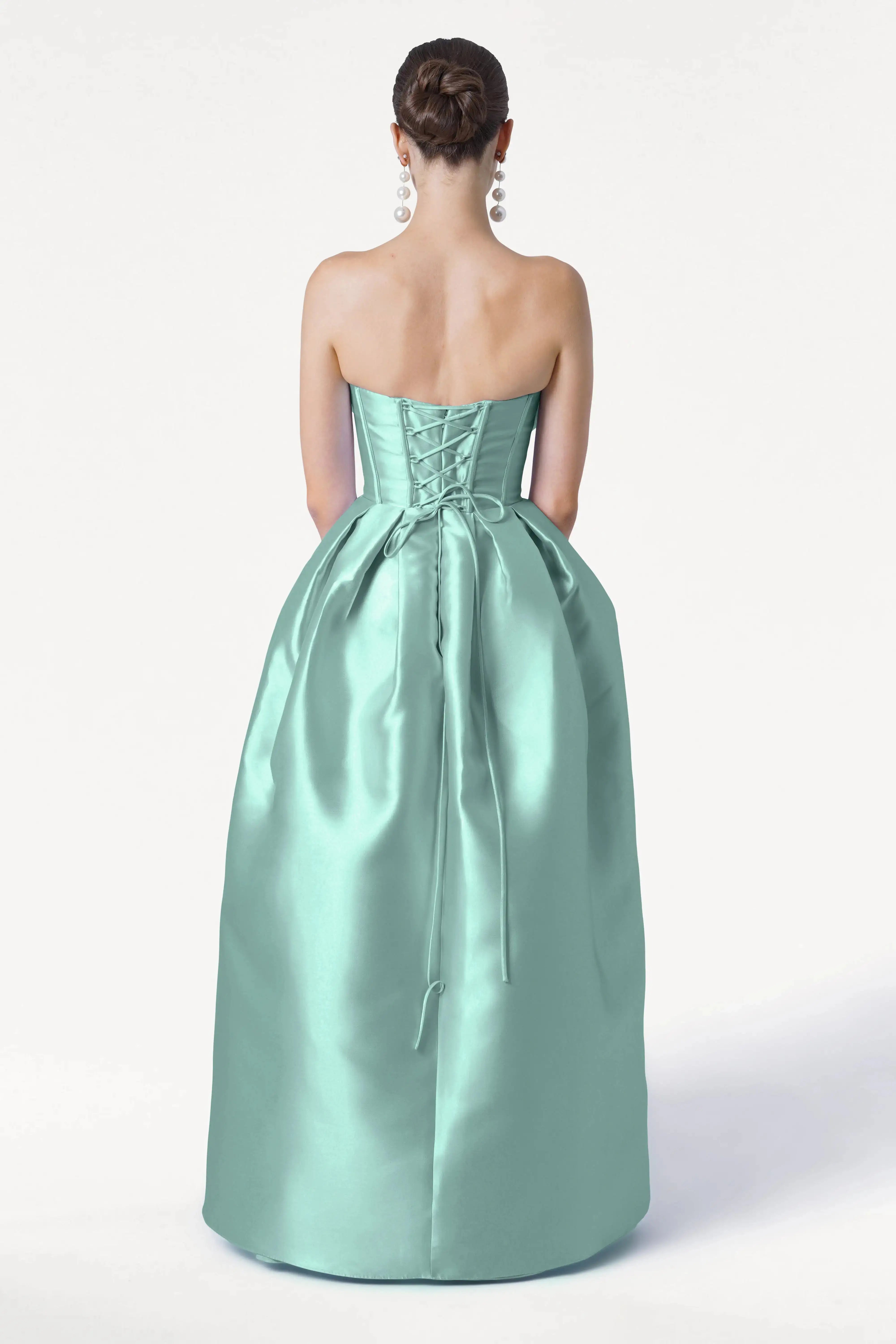 A woman with her hair in a bun and large earrings models the TH&TH Ophelia Bridesmaid Dress in Seafoam Satin Twill, featuring a strapless corset bodice and lace-up back, standing against a plain white background.