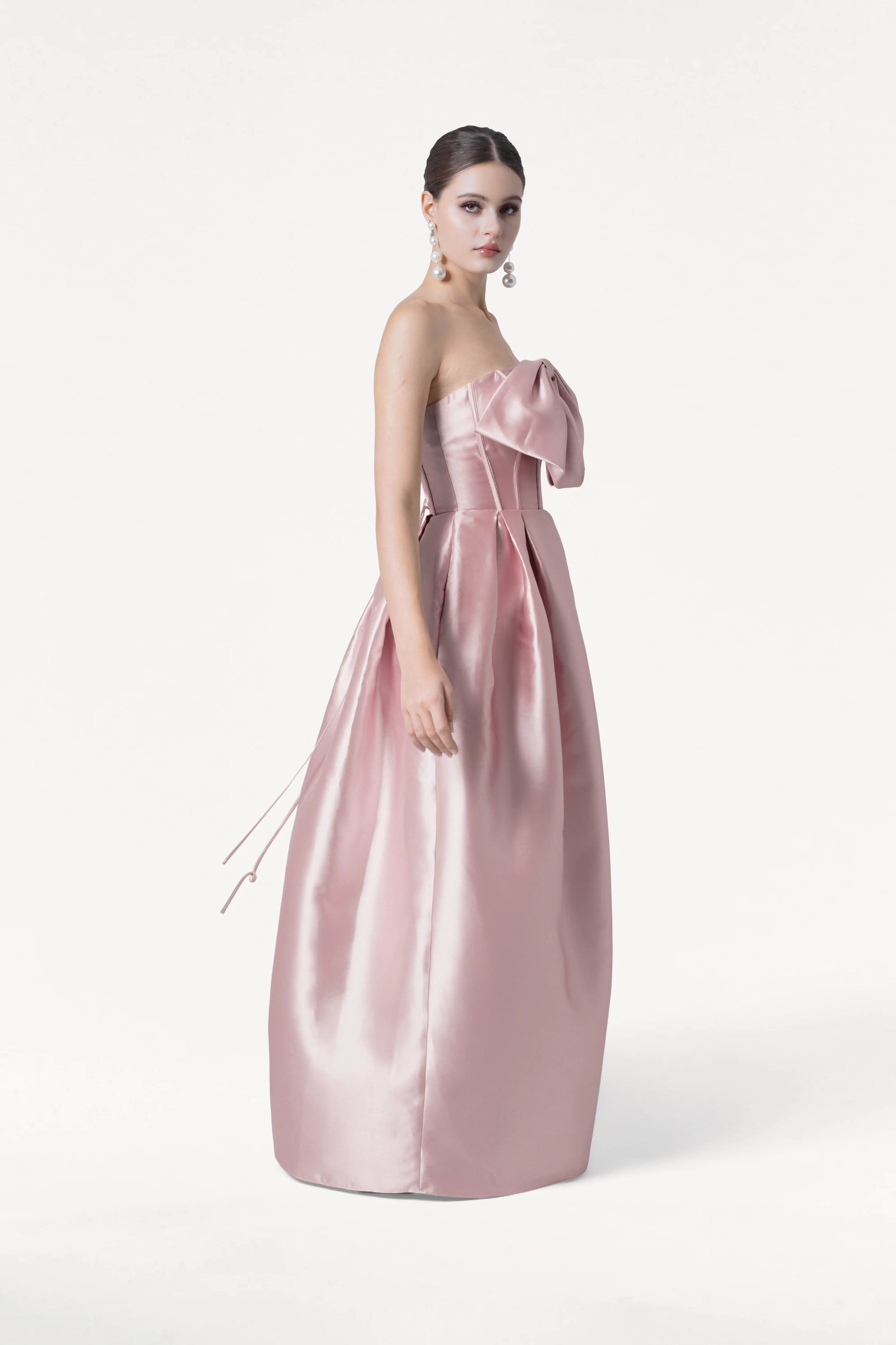 A woman poses against a white background in the TH&TH Ophelia Bridesmaid Dress, a strapless, floor-length smoked blush satin twill gown featuring a large bow on the corset bodice. She wears dangly earrings with her dark hair neatly styled back.