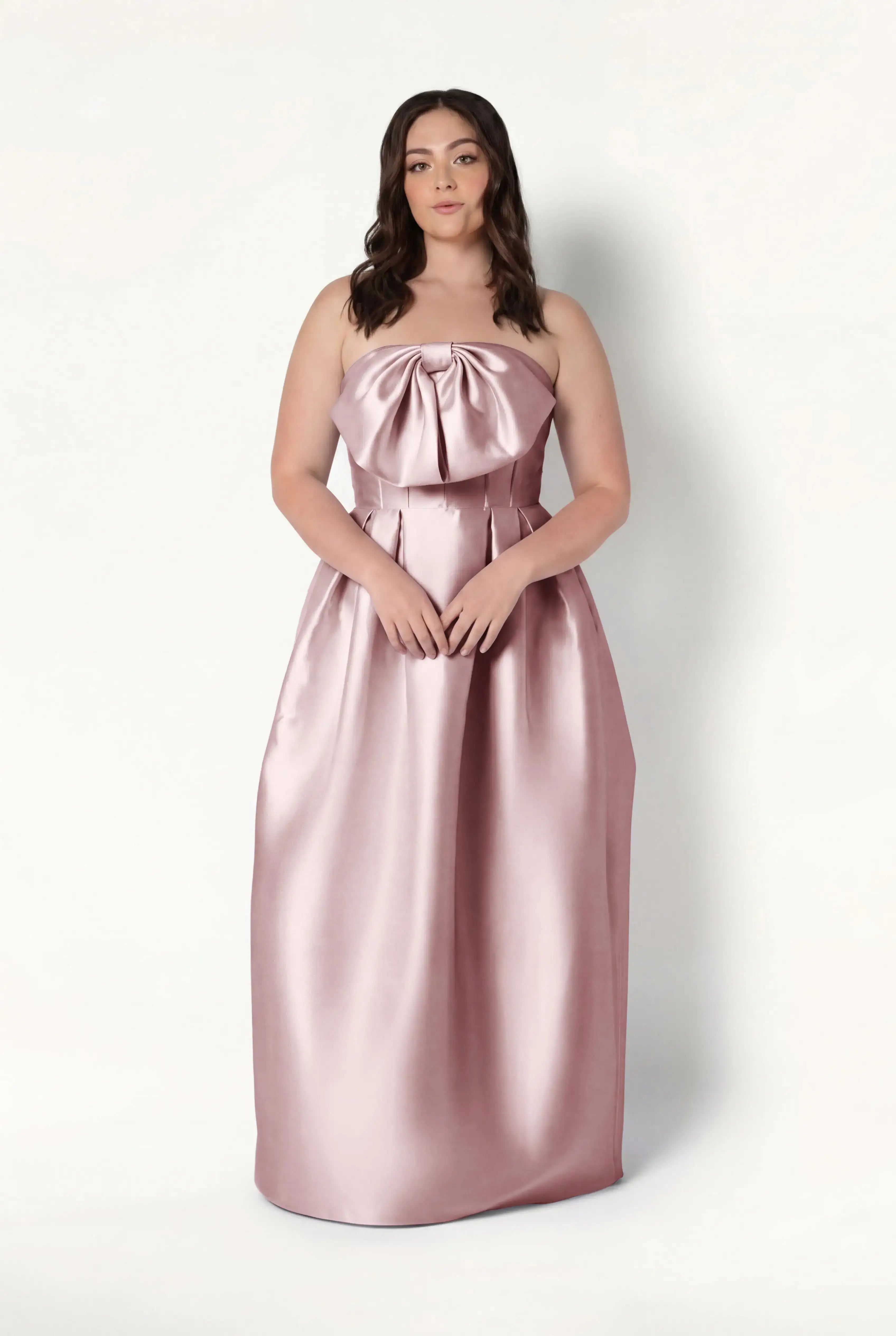 A woman with long brown hair wears the TH&TH Ophelia Bridesmaid Dress in smoked blush satin twill, featuring a strapless corset bodice and large bow, standing against a plain white background.