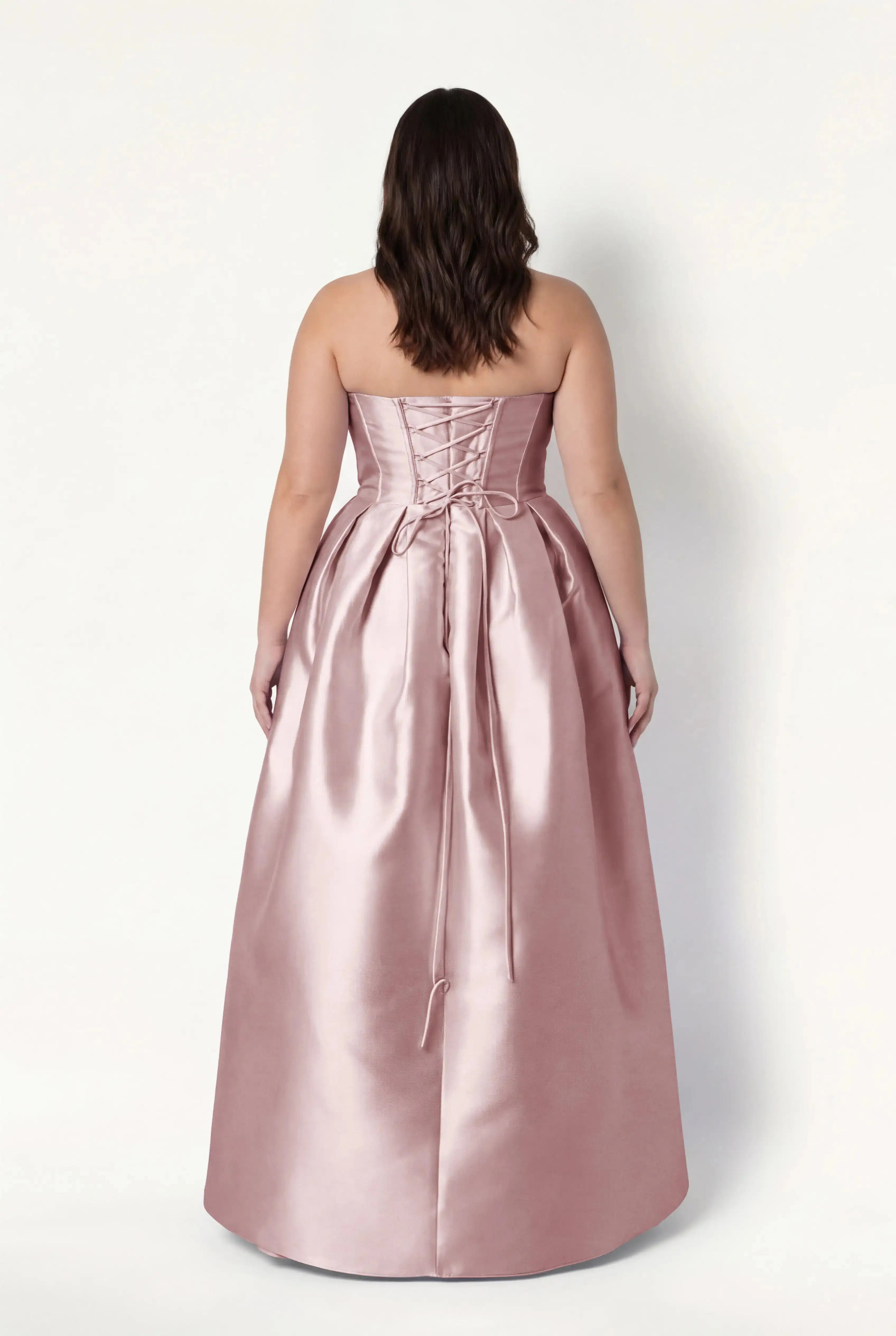 A woman with wavy brown hair wears the TH&TH Ophelia Bridesmaid Dress in Smoked Blush Satin Twill, featuring a strapless lace-up corset bodice, long ribbon ties, and floor-length skirt. She faces away against a plain white background.