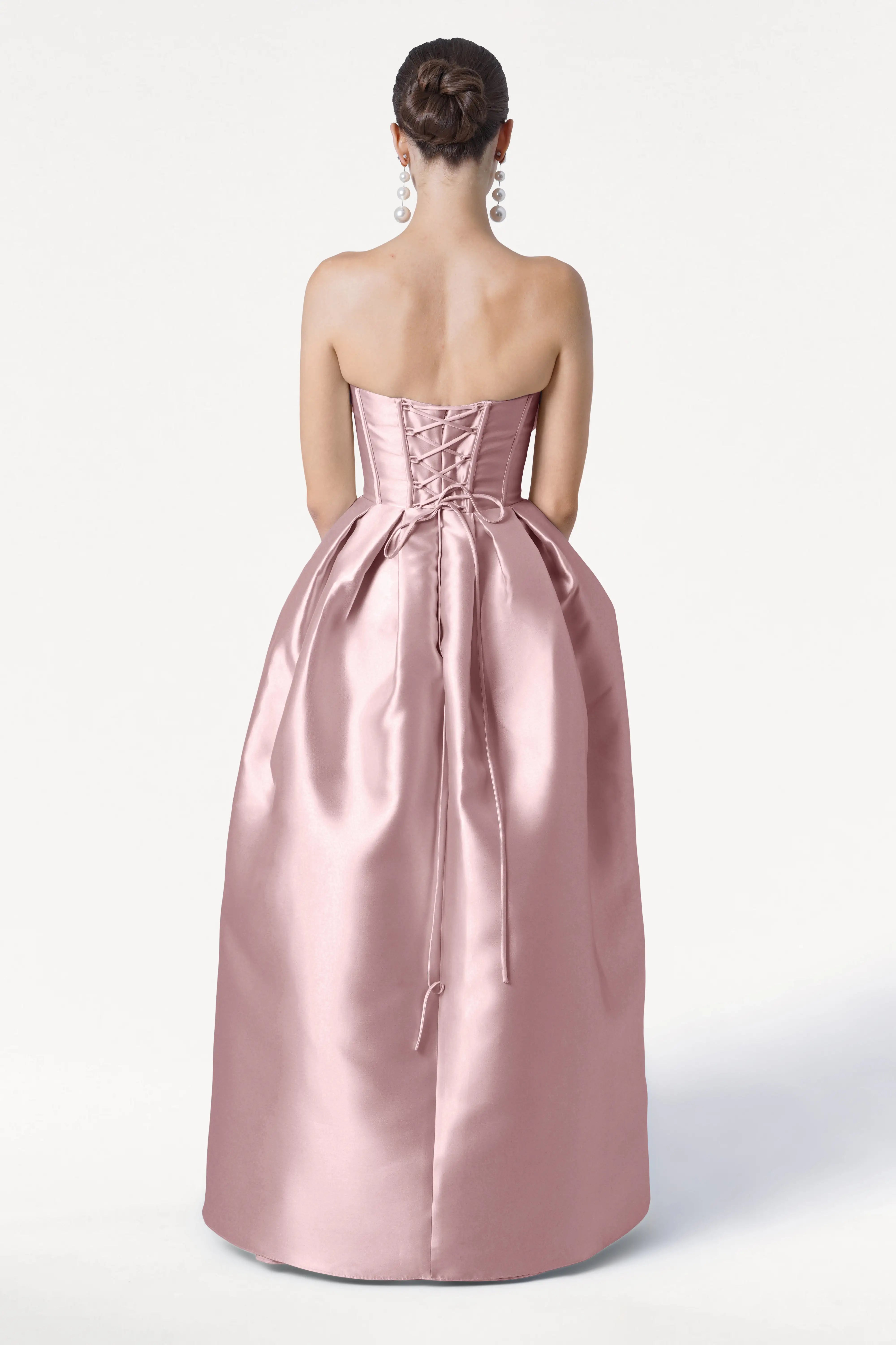 A woman with her hair in a bun and long earrings wears the TH&TH Ophelia Bridesmaid Dress in Smoked Blush Satin Twill, featuring a strapless corset bodice and lace-up back, standing against a plain white background.