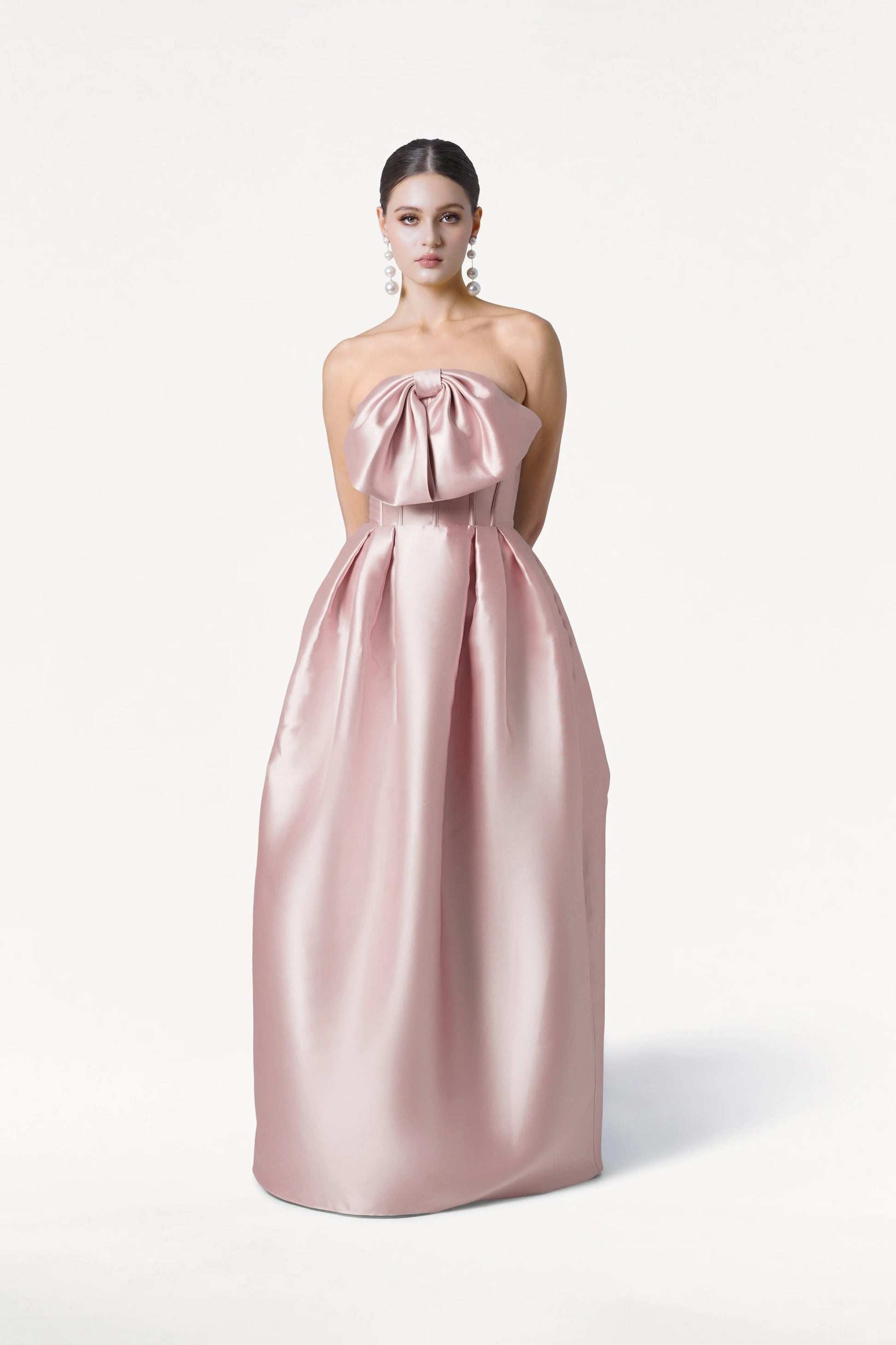 A woman stands against a white background in the TH&TH Ophelia Bridesmaid Dress in Smoked Blush Satin Twill, a strapless floor-length gown with a bow-shaped corset bodice and pleated skirt. Her hair is pulled back and she wears large pearl earrings.