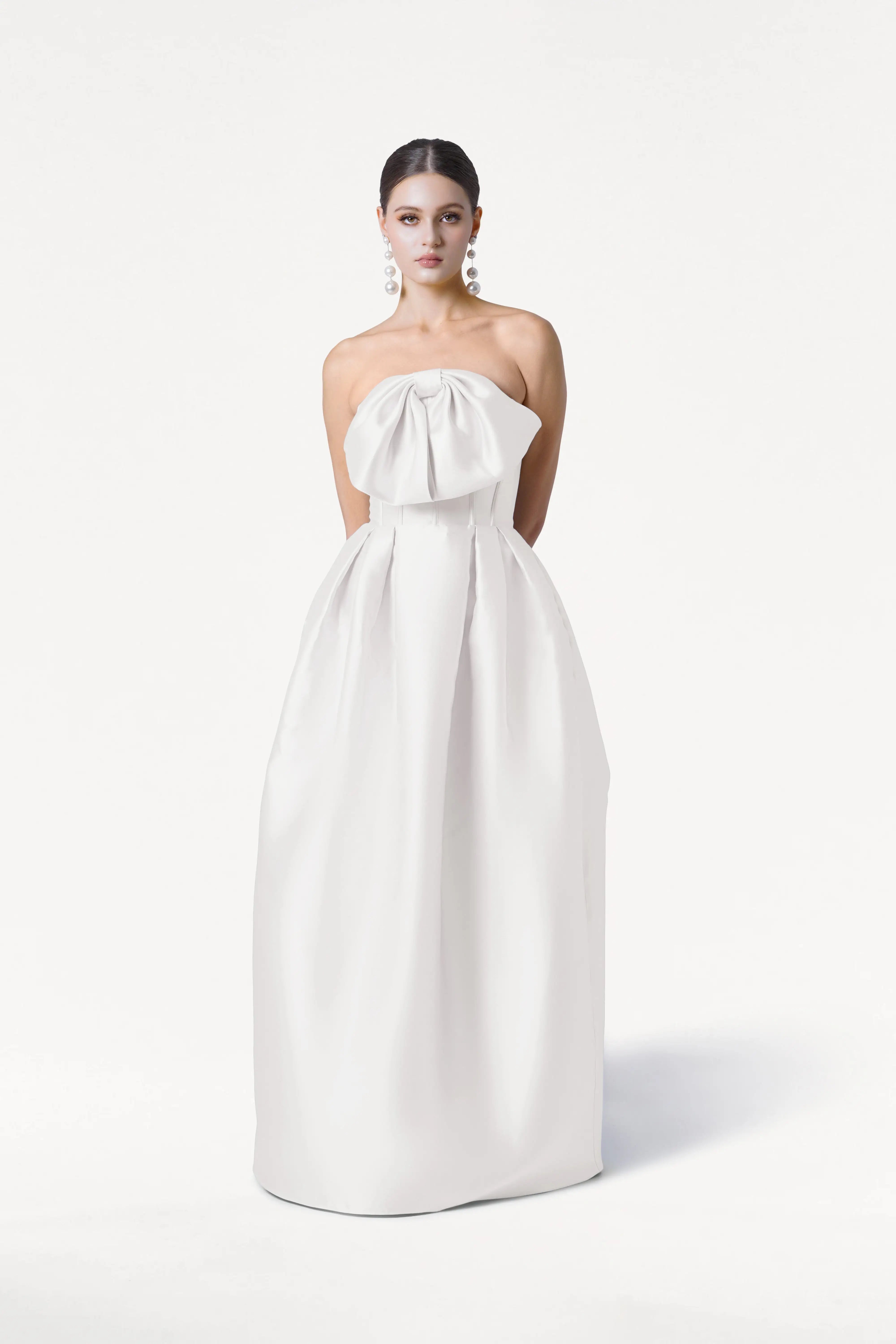 A woman stands against a white background wearing the TH&TH Ophelia Dress in Ivory Satin Twill, featuring a strapless neckline, large bow detail, and full floor-length skirt. She pairs it with pearl drop earrings and has her hair styled back.