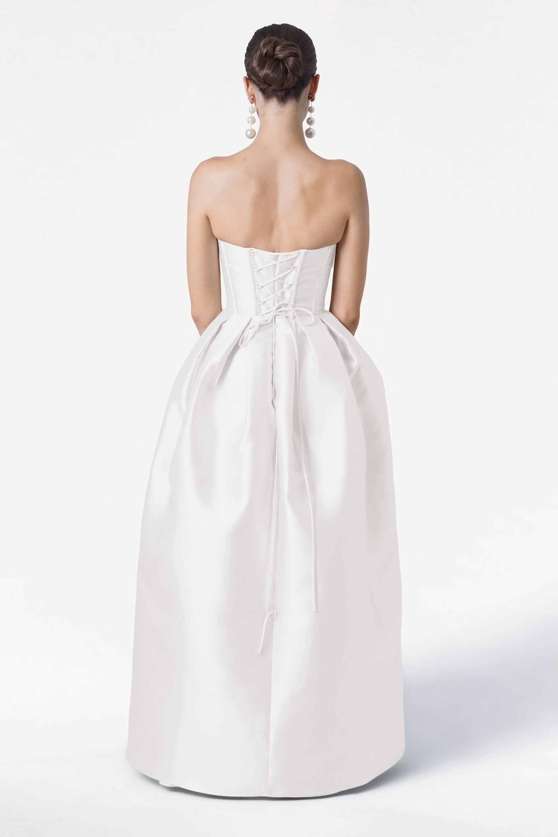 A woman with dark hair in a bun faces away from the camera against a white background, wearing the TH&TH Ophelia Dress in Ivory Satin Twill—a strapless gown with a corset bodice and full skirt—and dangling silver earrings.
