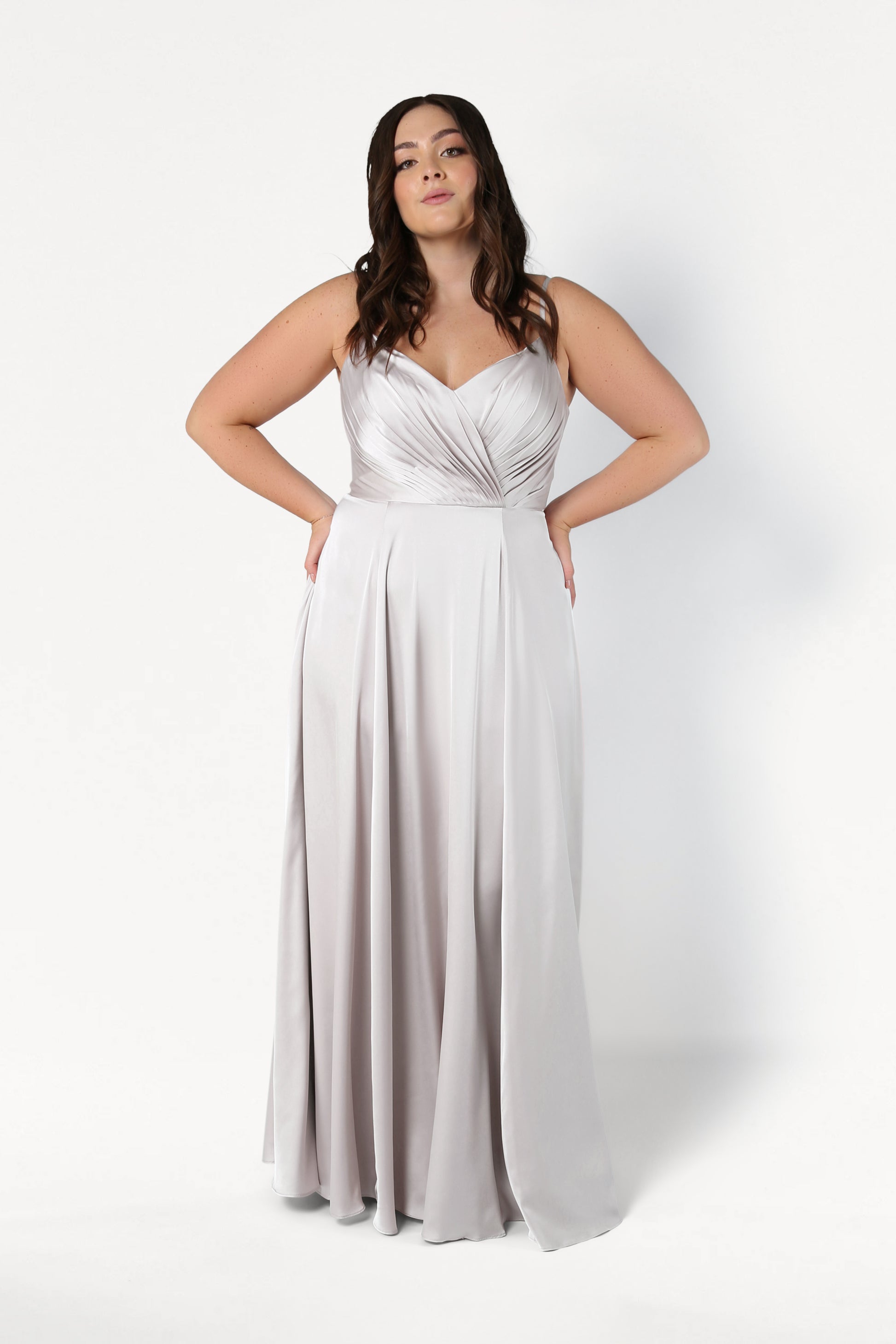 A woman stands against a white background wearing the TH&TH Isla Bridesmaid Dress in Oyster Satin, featuring a pleated bodice, thin straps, flowing skirt, and pockets. Her long, wavy dark hair cascades down.