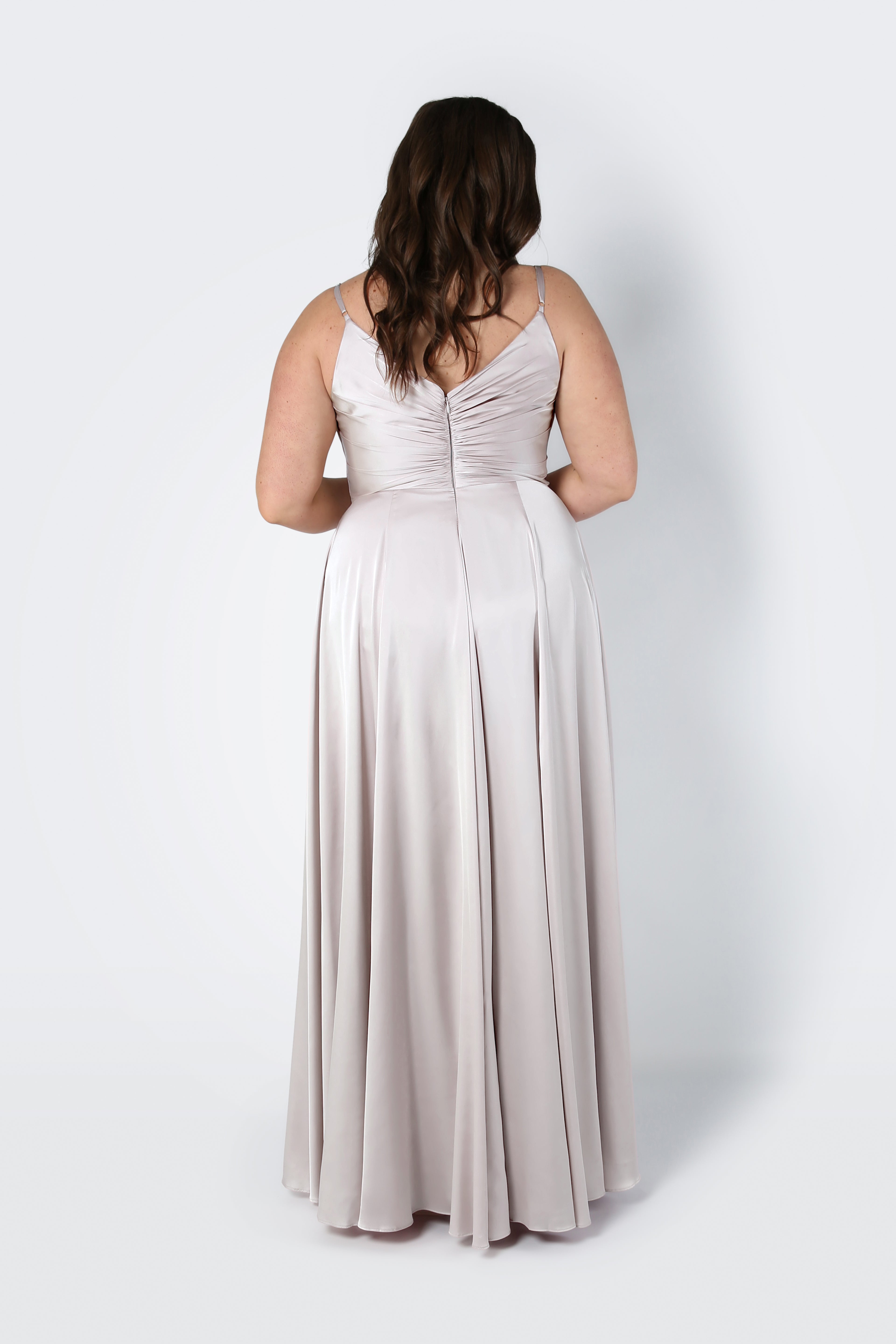 A woman with long brown hair stands facing away, wearing the TH&TH Isla Bridesmaid Dress in Oyster Satin—featuring thin straps, a ruched back, and a pleated bodice—against a plain white background.