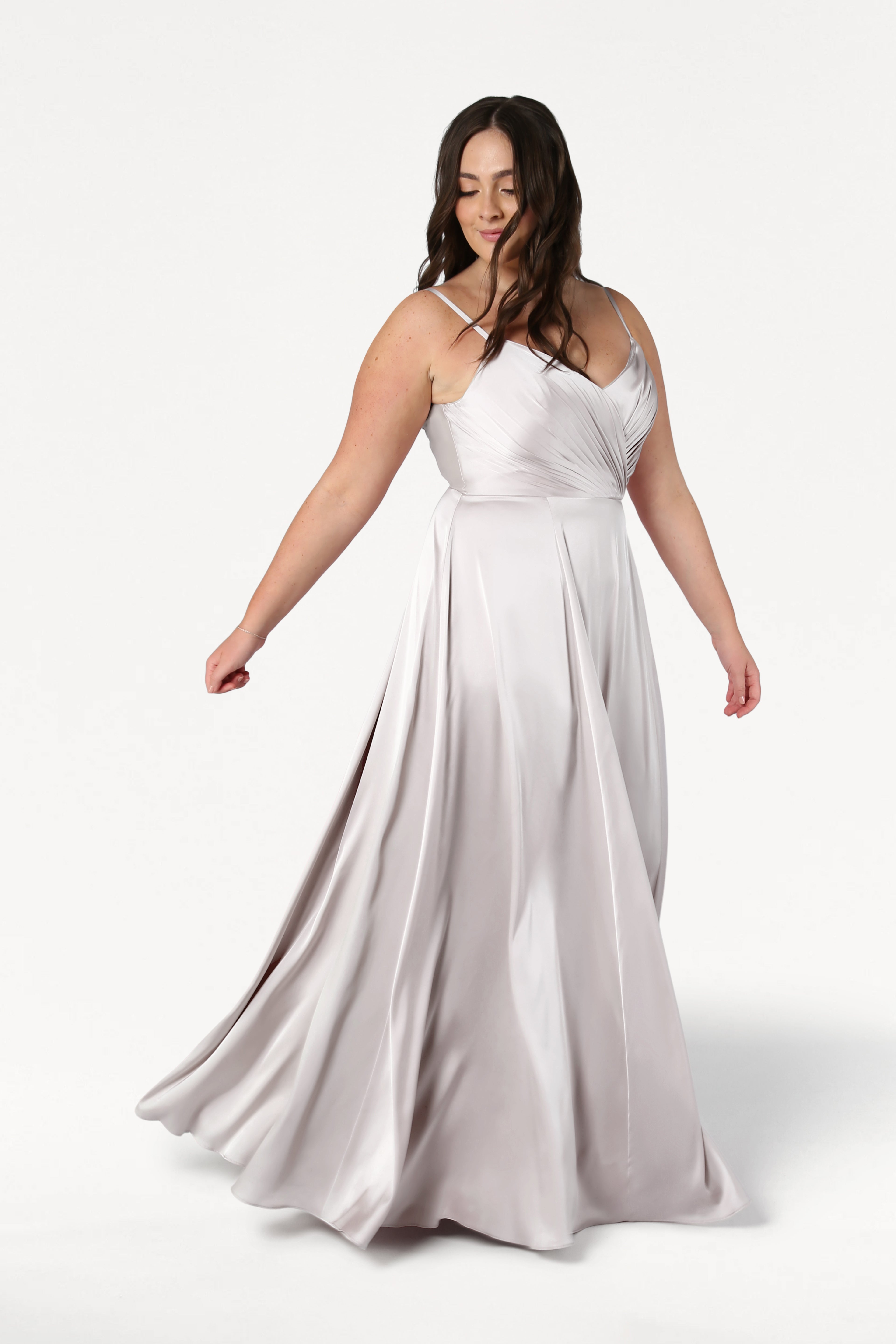A woman twirls gracefully against a white background in the Isla Bridesmaid Dress in Oyster Satin by TH&TH, featuring a flowing silhouette, satin pleated bodice, and delicate spaghetti straps.