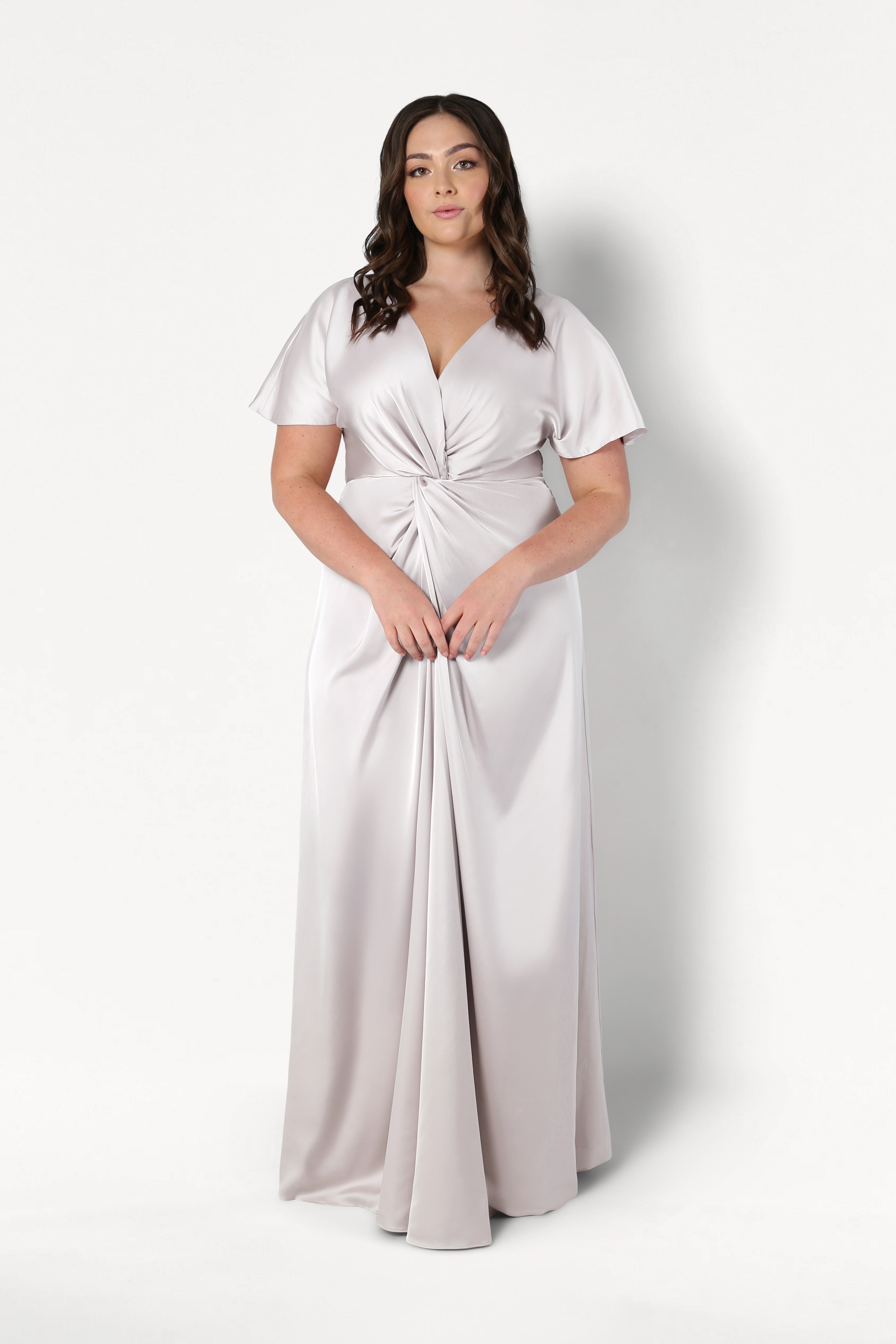 A woman with long brown hair wears the TH&TH Camilla Bridesmaid Dress in Oyster Satin, featuring short flutter sleeves and a twist waist detail, standing against a plain white background.