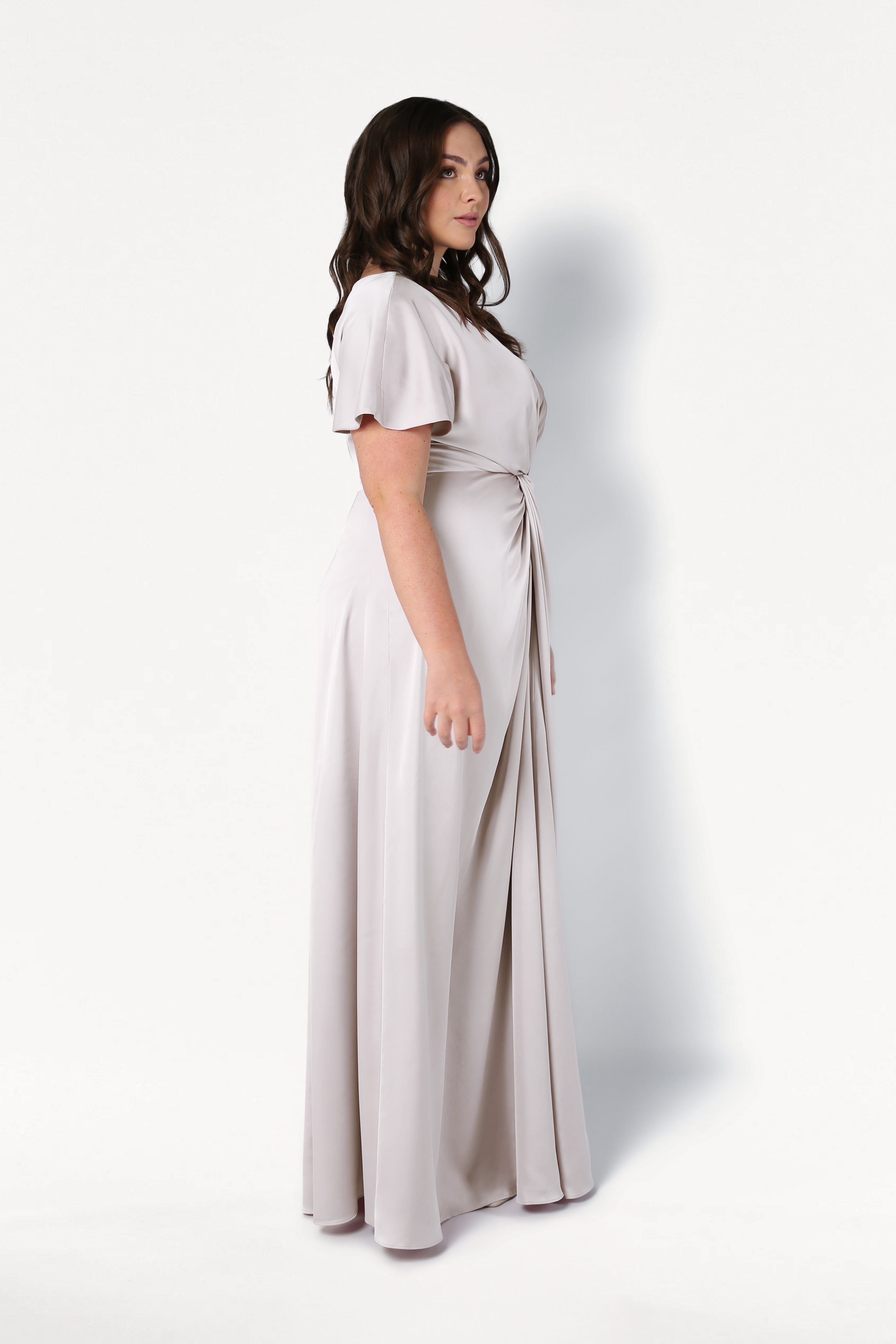 A woman with long dark hair stands in profile against a white background, wearing the TH&TH Camilla Bridesmaid Dress in Oyster Satin—a size-inclusive, floor-length gown featuring short sleeves and a gathered waist.