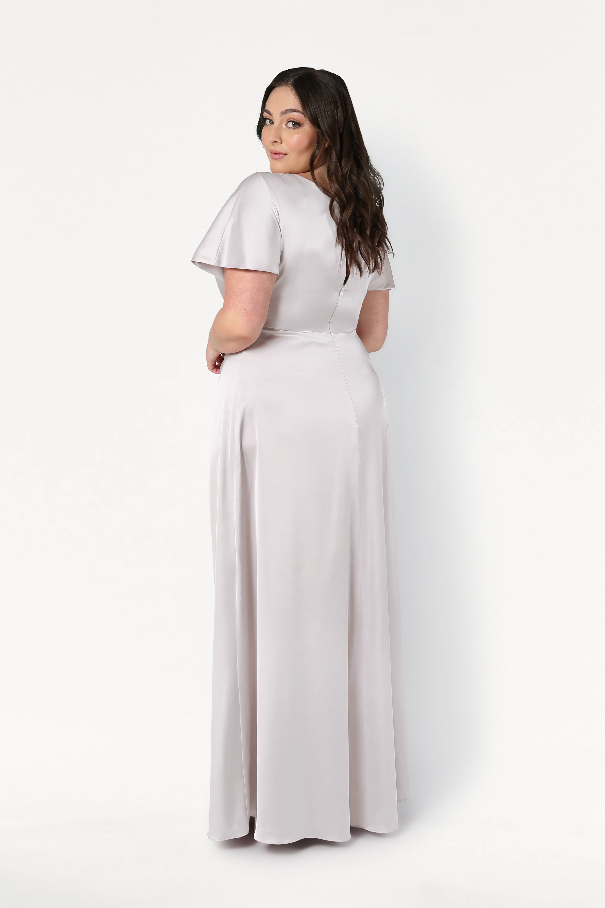 A woman with long dark hair wears the TH&TH Camilla Bridesmaid Dress in Oyster Satin, floor-length and light gray. She faces away, looking over her shoulder with a neutral expression against a plain white background.