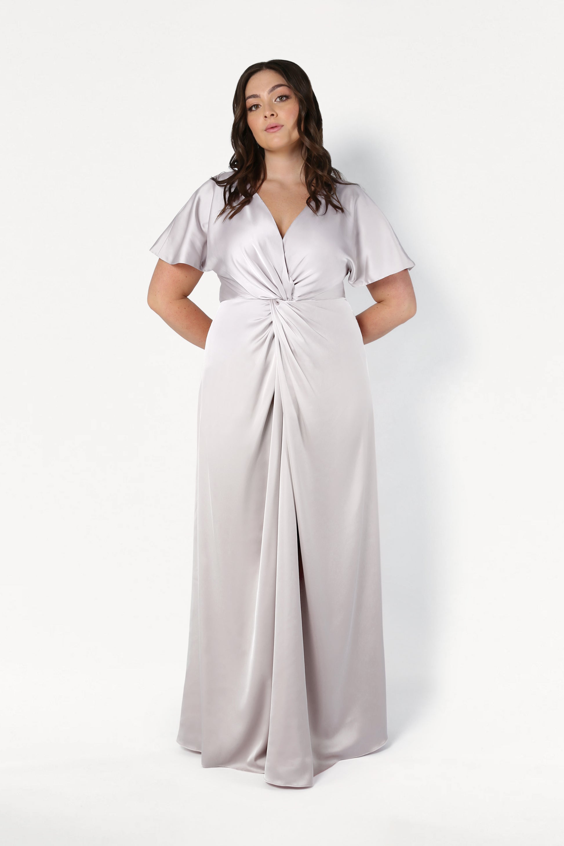 A woman stands in front of a plain white background wearing the TH&TH Camilla Bridesmaid Dress in Oyster Satin, featuring short flutter sleeves, a V-neckline, and gathered twist detail at the waist.