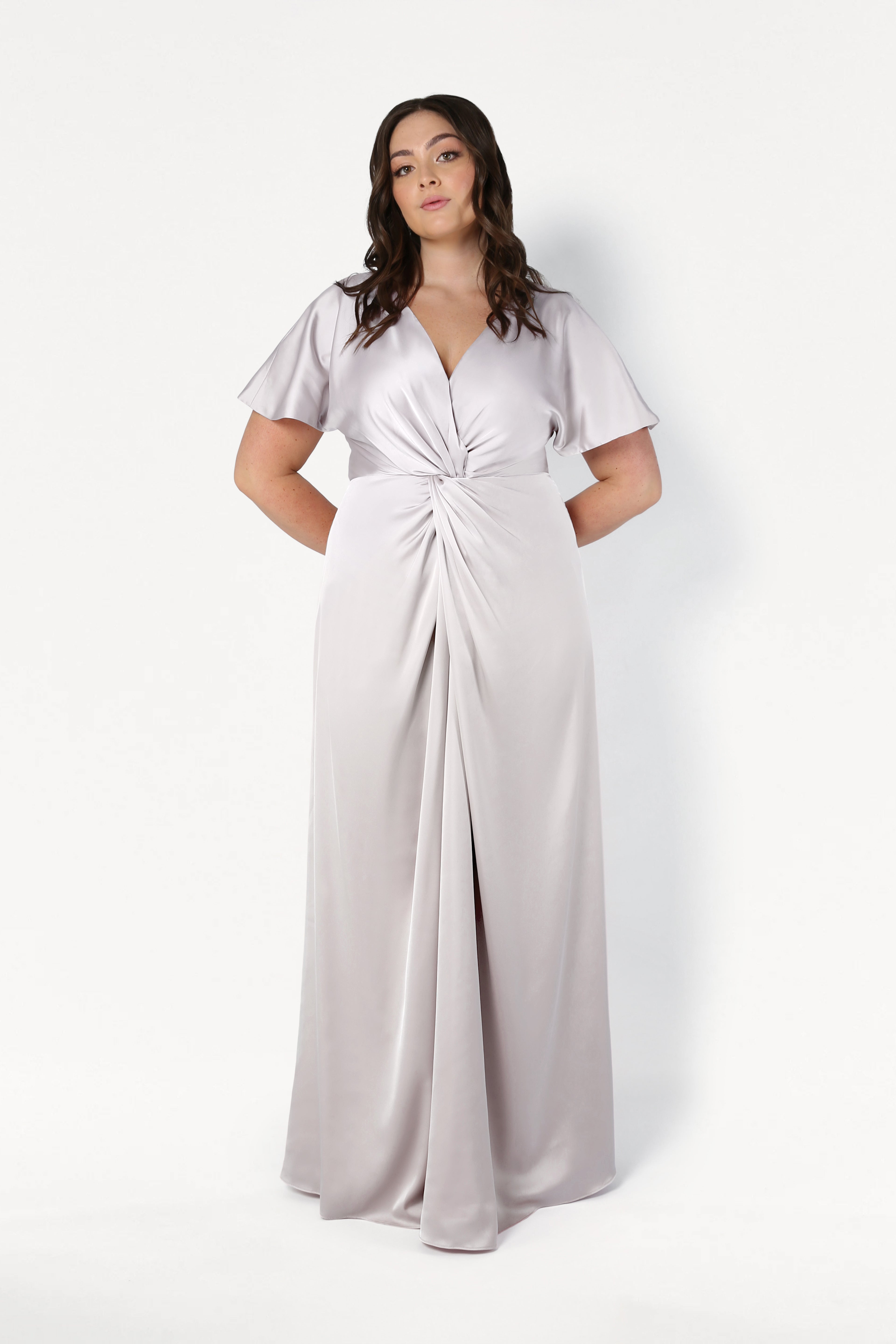 A woman stands in front of a plain white background wearing the TH&TH Camilla Bridesmaid Dress in Oyster Satin, featuring short flutter sleeves, a V-neckline, and gathered twist detail at the waist.