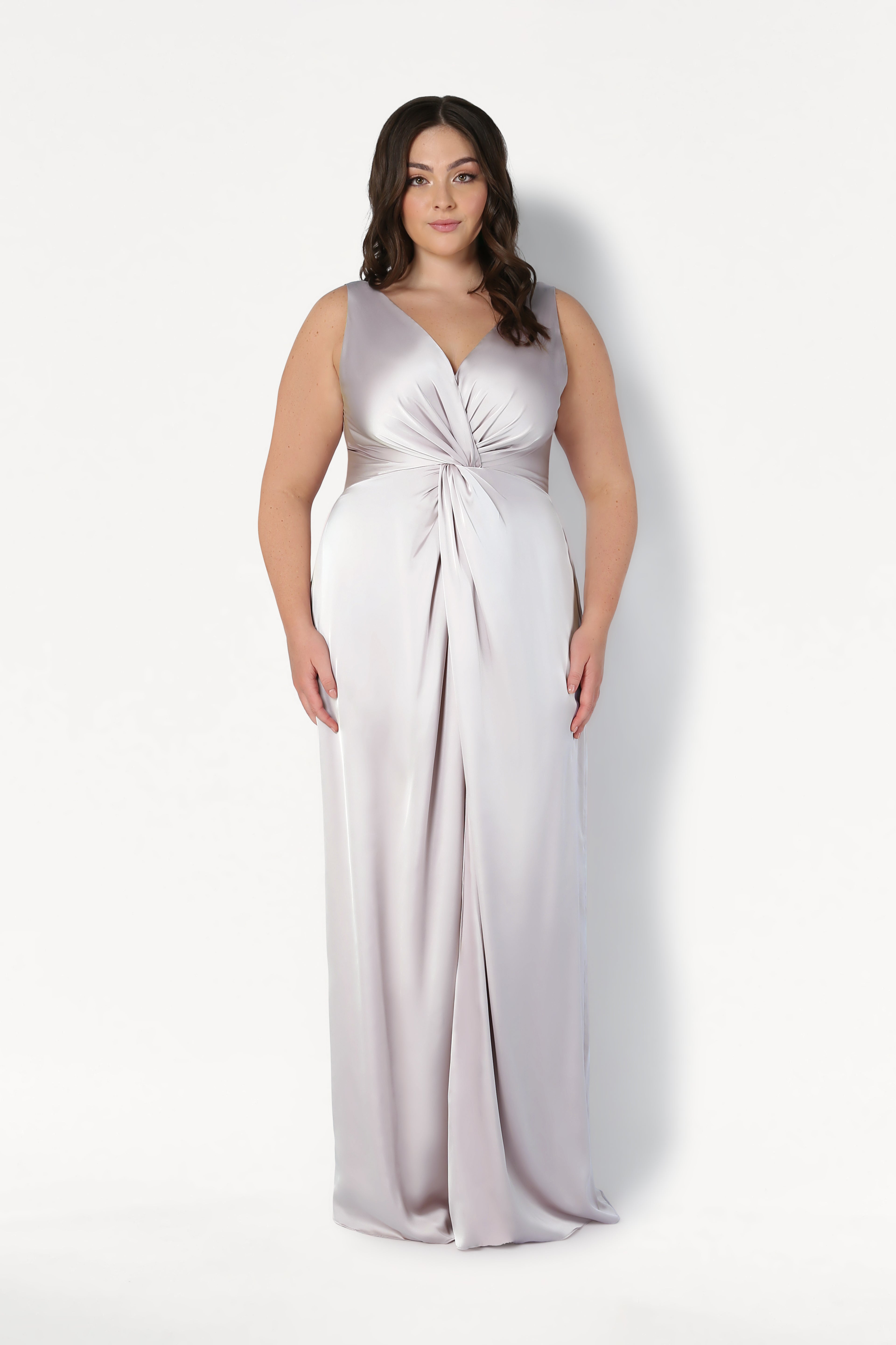 A woman stands against a white background in the TH&TH Carmen Bridesmaid Dress in Oyster Satin, a sleeveless, floor-length gown with a twist-knot front—ideal for modern bridal parties. She wears her long dark hair in loose waves and has a neutral expression.