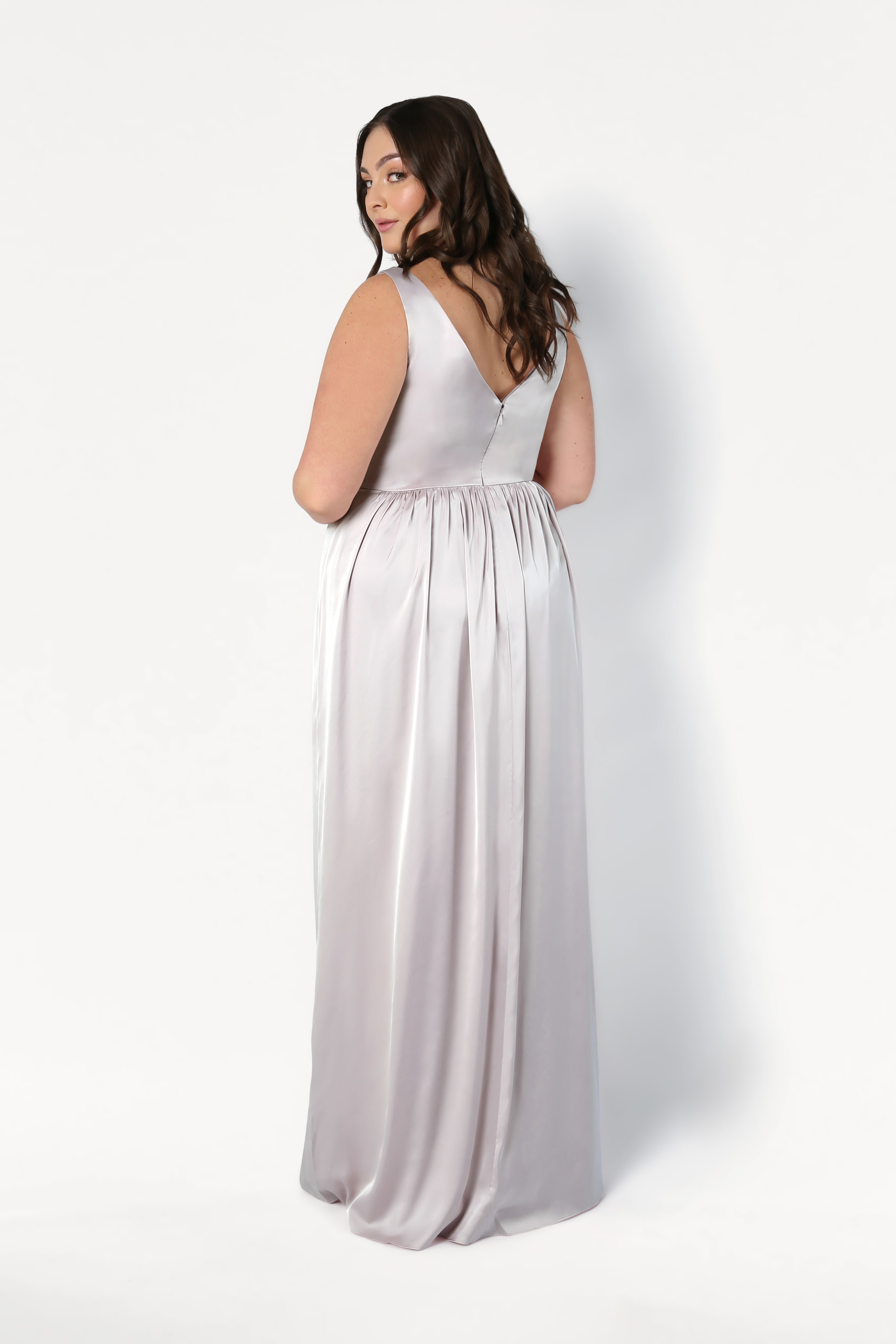 A woman in the TH&TH Carmen Bridesmaid Dress in Oyster Satin, a sleeveless, floor-length gown with a twist-knot front, stands facing away and looks over her shoulder against a plain white background.