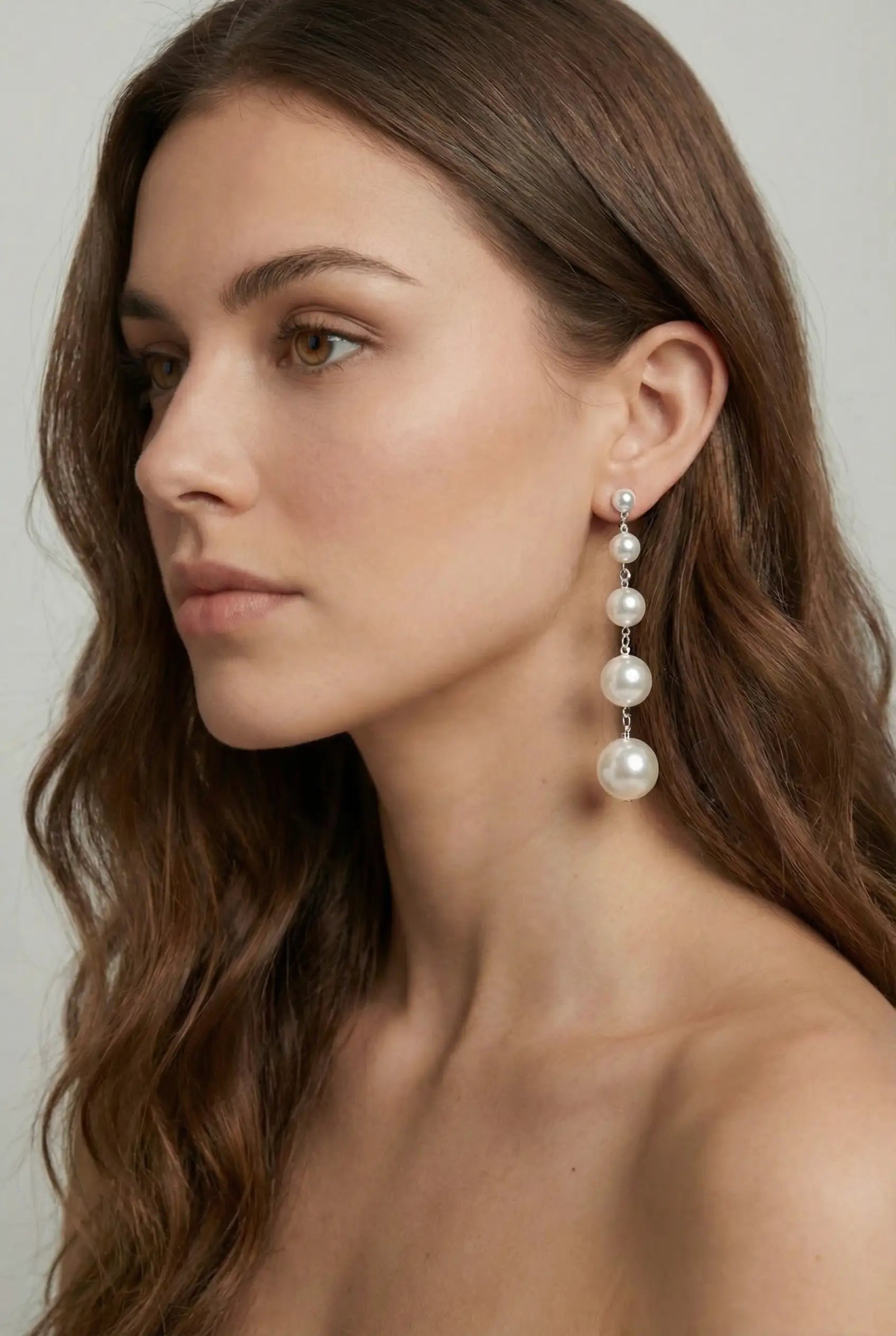 A woman with long brown hair models the TH&TH Palais Pearl Drop Earrings, featuring elegant white pearls in varying sizes. She faces left in a bright, neutral setting—an ideal scene for showcasing these bridal earrings.