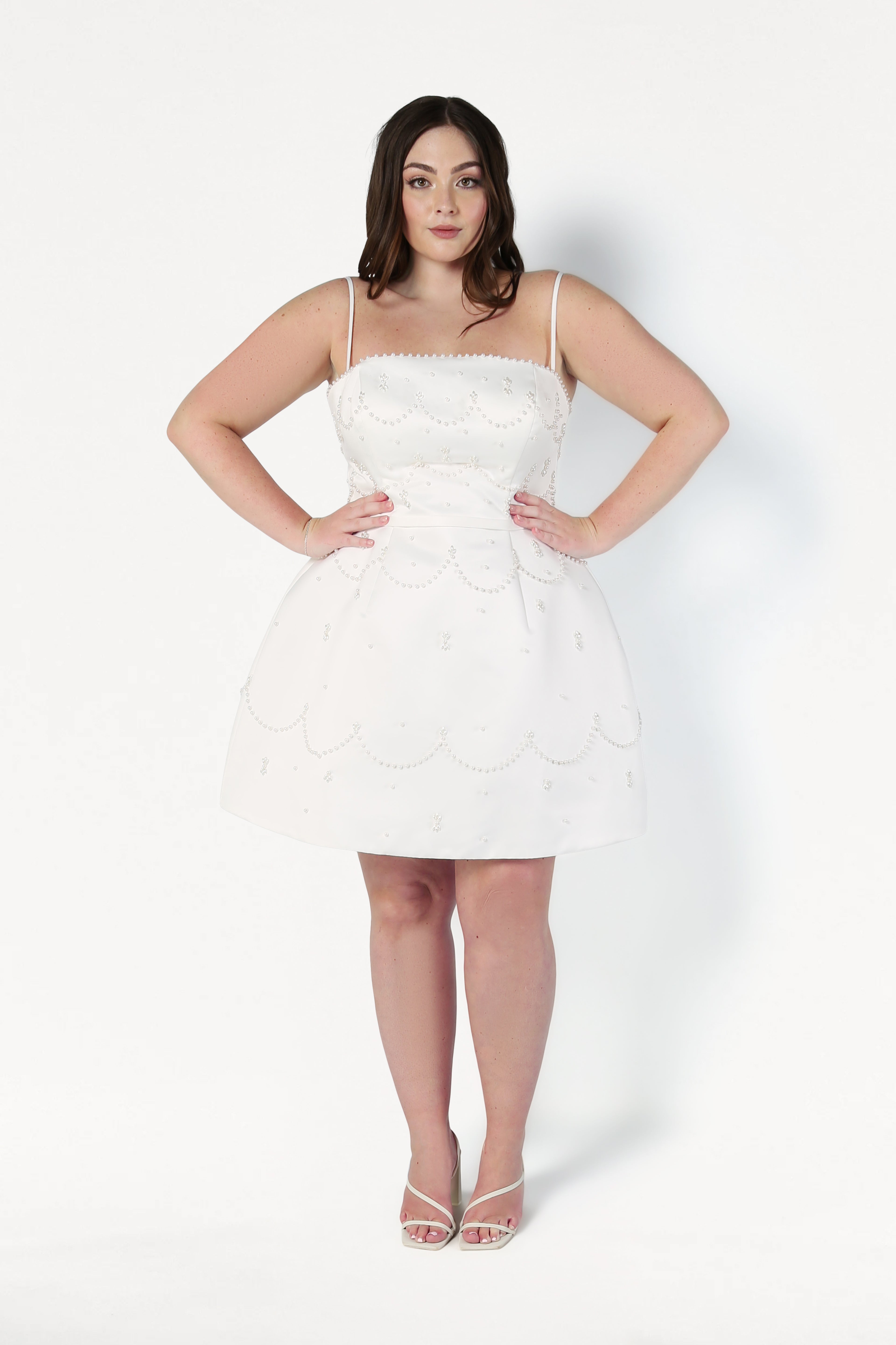 A woman with long dark hair wears the TH&TH Pearl Bridal Mini Dress in Ivory, a sleeveless mini dress with scalloped edges and pearl detailing, standing facing forward against a white background with hands on hips and open-toed heels.
