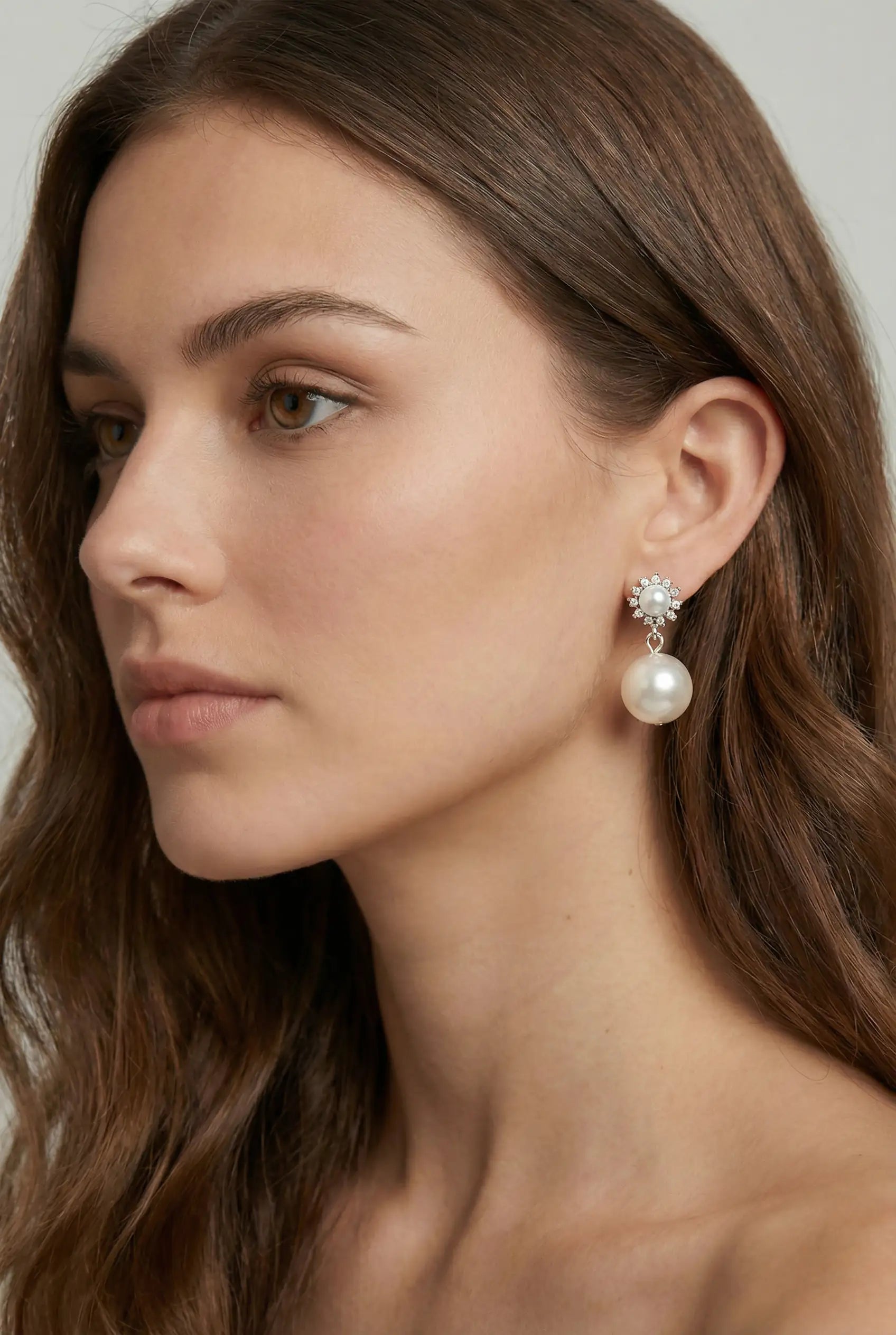 A woman with long brown hair and natural makeup wears TH&TH Pearl Blossom Stud Earrings—elegant bridal earrings with pearls and crystals—in a close-up side profile against a neutral background.