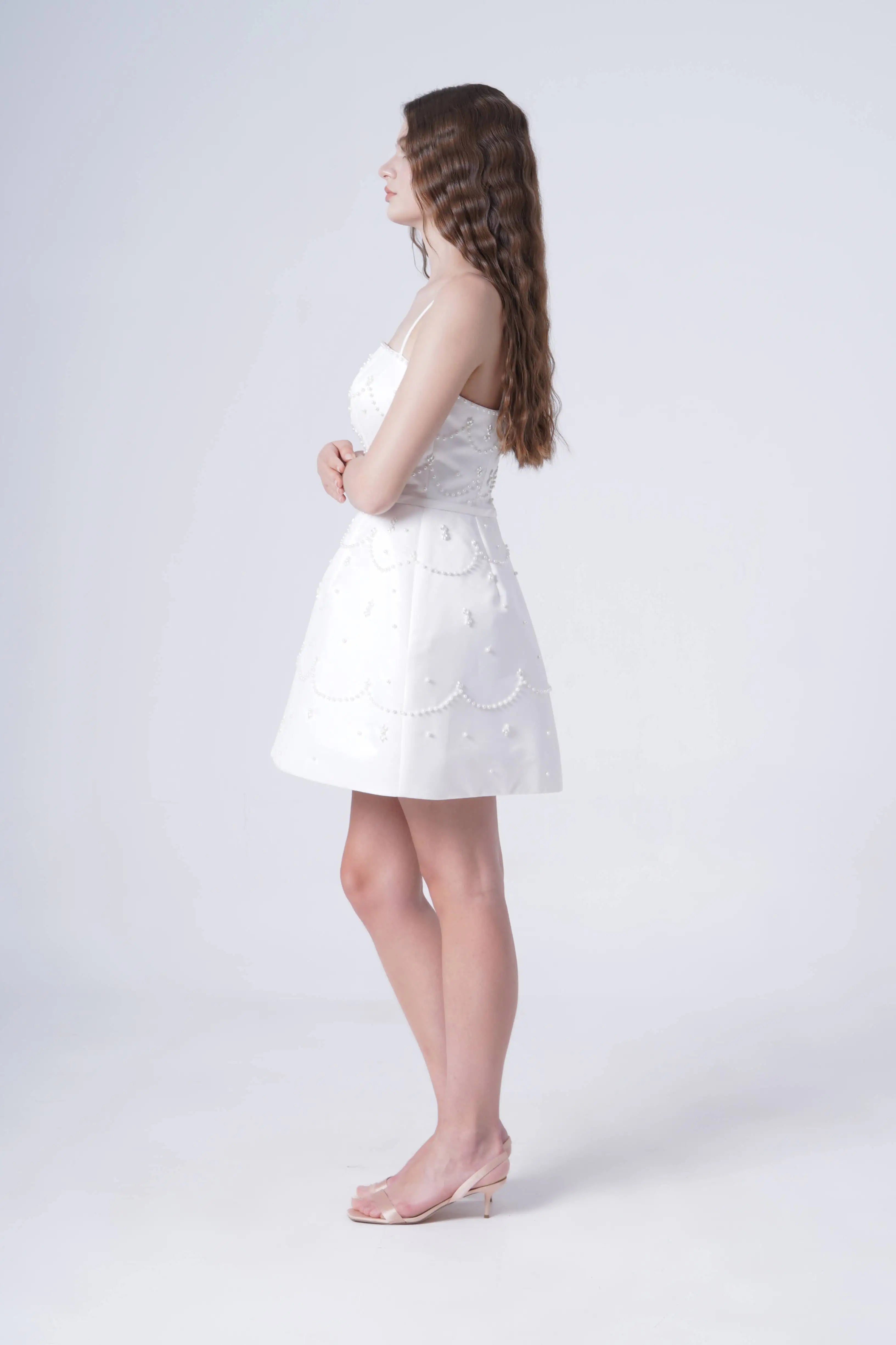 A woman with long, wavy brown hair stands in profile against a white background, wearing the TH&TH Pearl Bridal Mini Dress in Ivory and translucent heels, her arms gently folded at her waist.