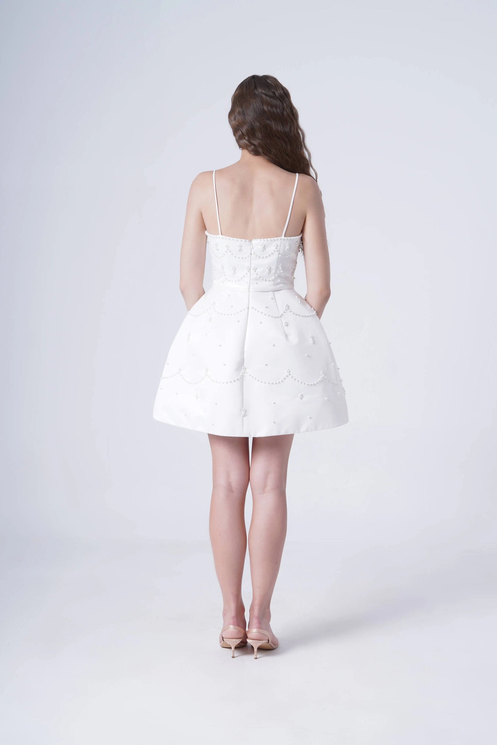 A woman faces away, wearing the TH&TH Pearl Bridal Mini Dress in Ivory. The white sleeveless dress features thin straps, delicate pearl detailing, decorative patterns, and she pairs it with nude heels against a plain light background.