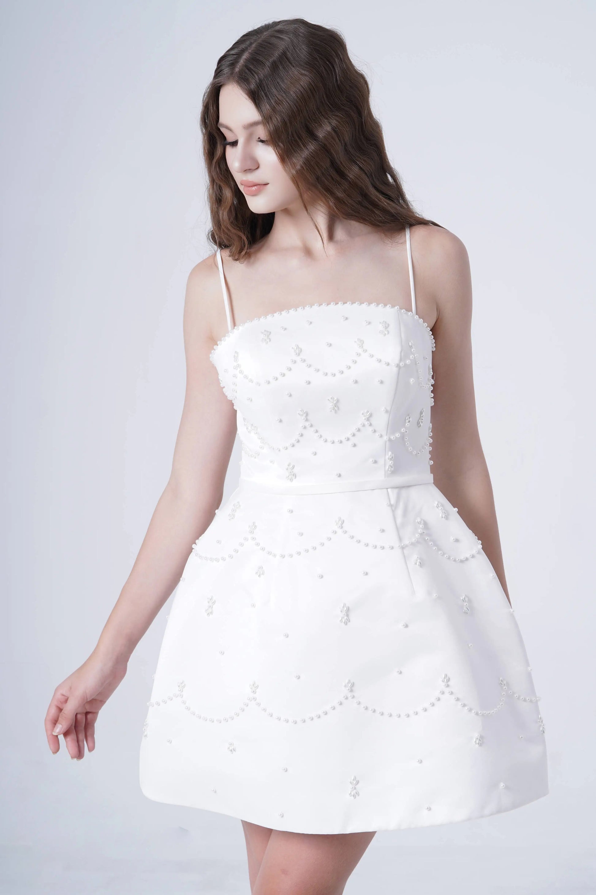 A woman with long wavy brown hair models the TH&TH Pearl Bridal Mini Dress in Ivory, featuring pearl details and a flared skirt, against a plain light background, gazing slightly to the side.