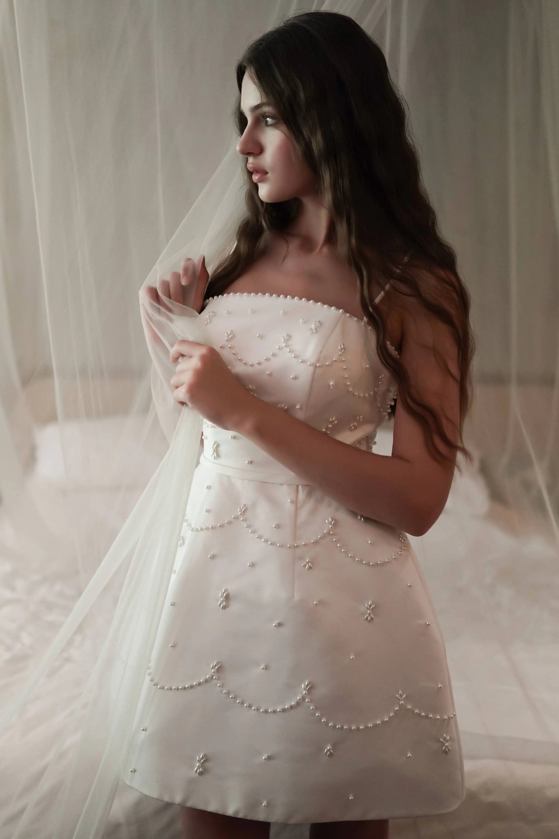 A young woman with long wavy hair stands indoors, wearing the TH&TH Pearl Bridal Mini Dress in Ivory and holding a sheer white veil, surrounded by soft lighting and delicate fabric drapery.