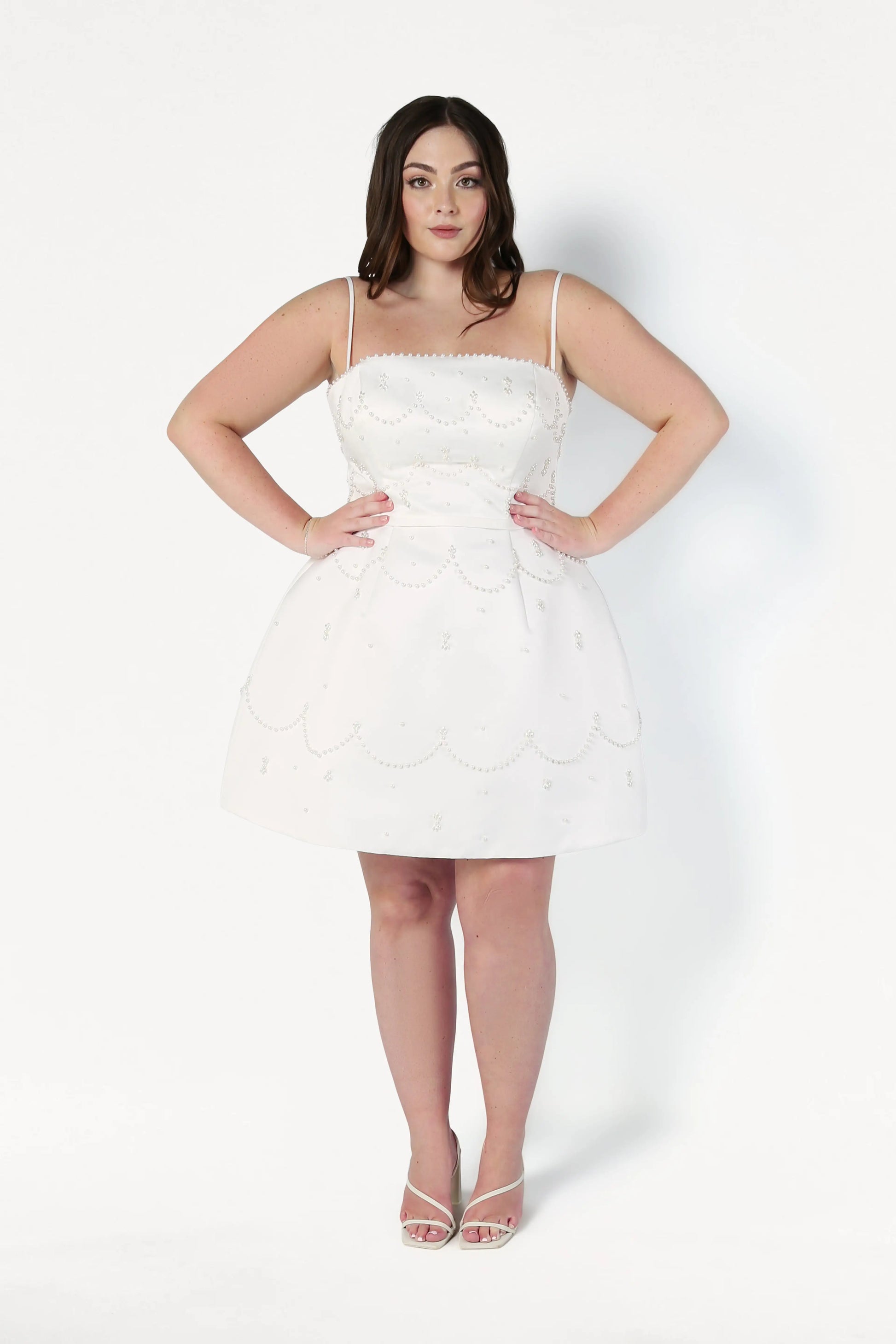 A woman with long dark hair wears the TH&TH Pearl Bridal Mini Dress in Ivory, a sleeveless mini dress with scalloped edges and pearl detailing, standing facing forward against a white background with hands on hips and open-toed heels.