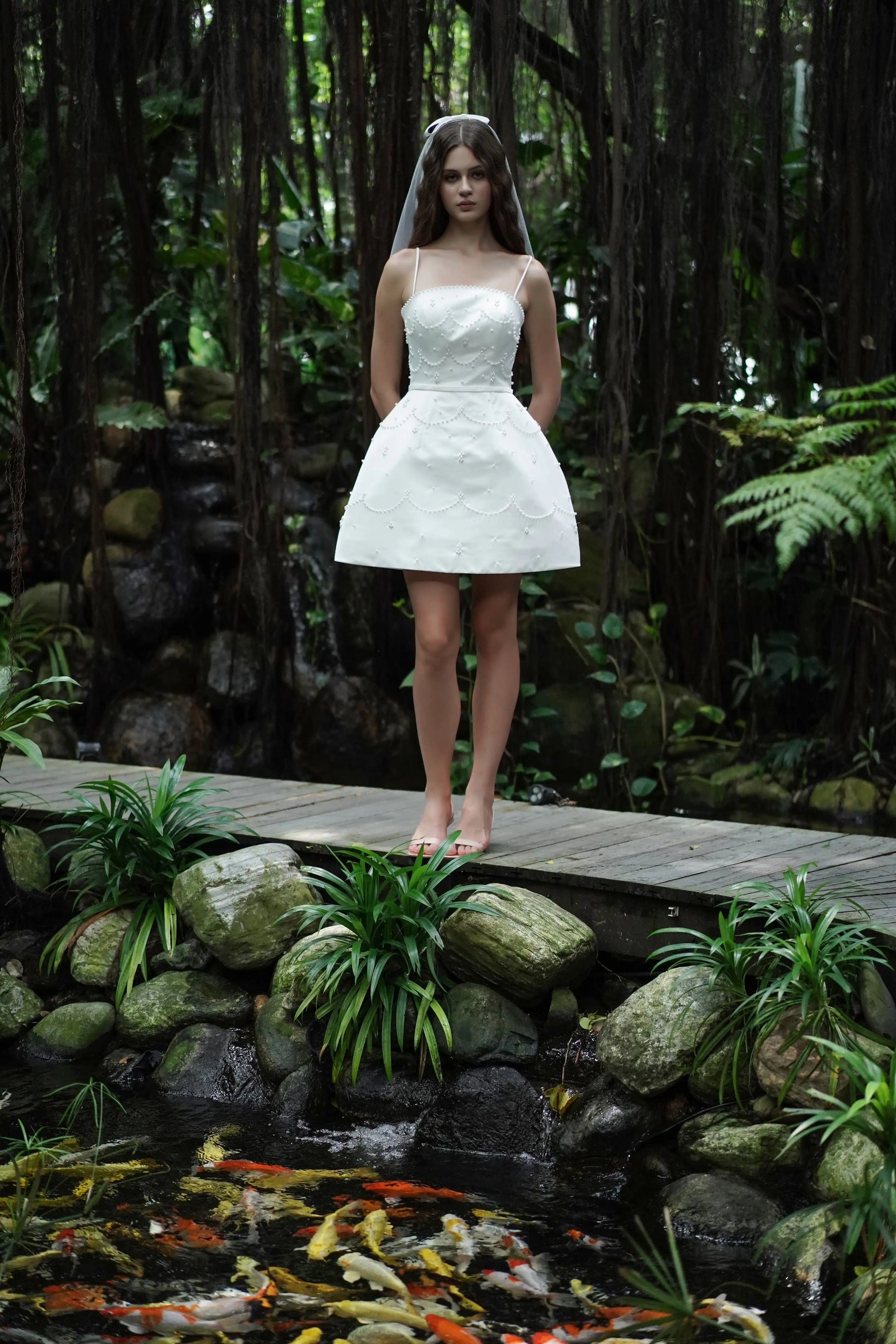 A woman wears the TH&TH Pearl Bridal Mini Dress in Ivory, standing barefoot on a wooden bridge in a lush garden beside rocks, greenery, and a pond with orange koi fish.