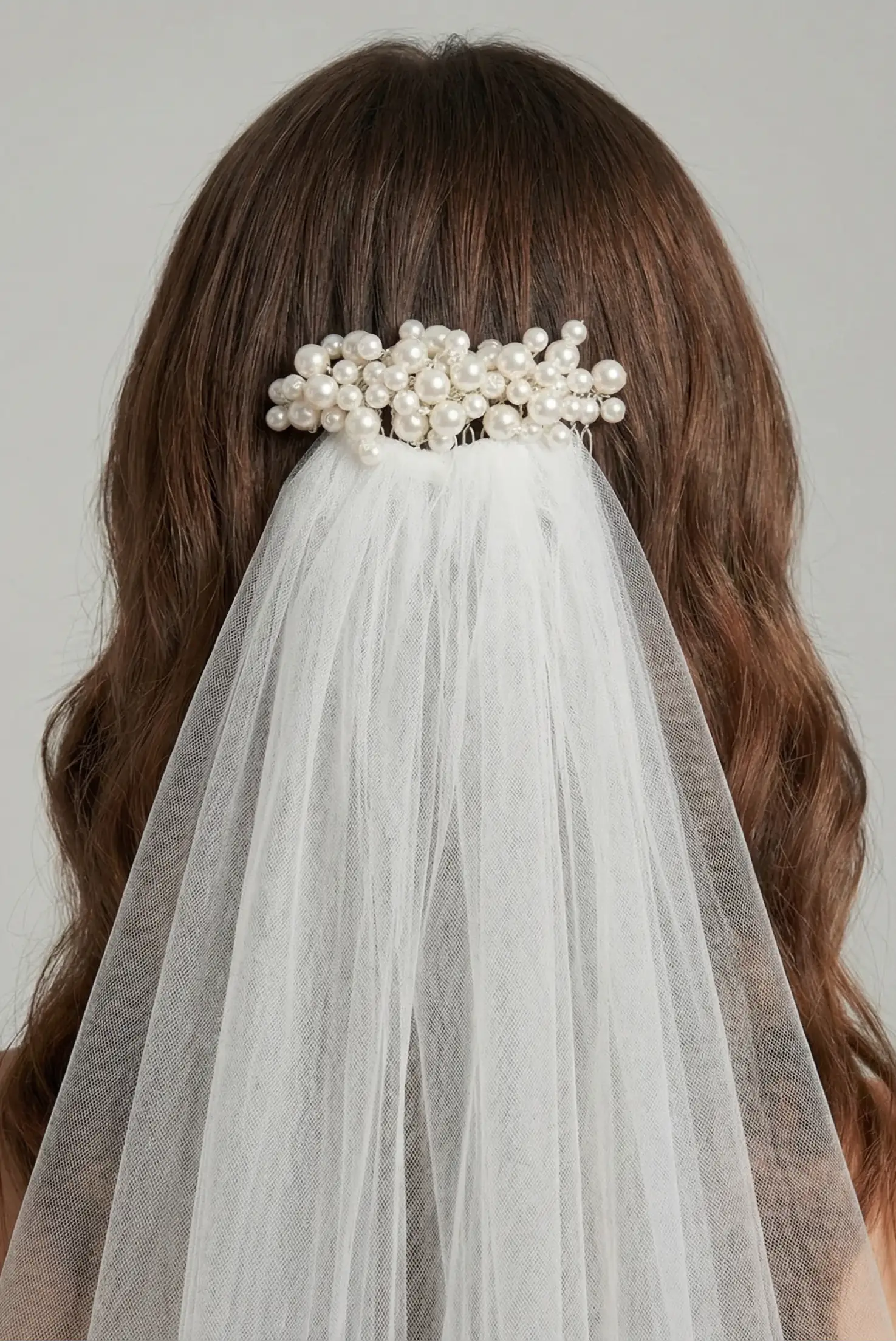 A woman with long brown wavy hair wears the TH&TH Pearl Cluster Hair Comb, which features clusters of white pearls securing a white tulle veil. She is photographed from behind against a plain background.
