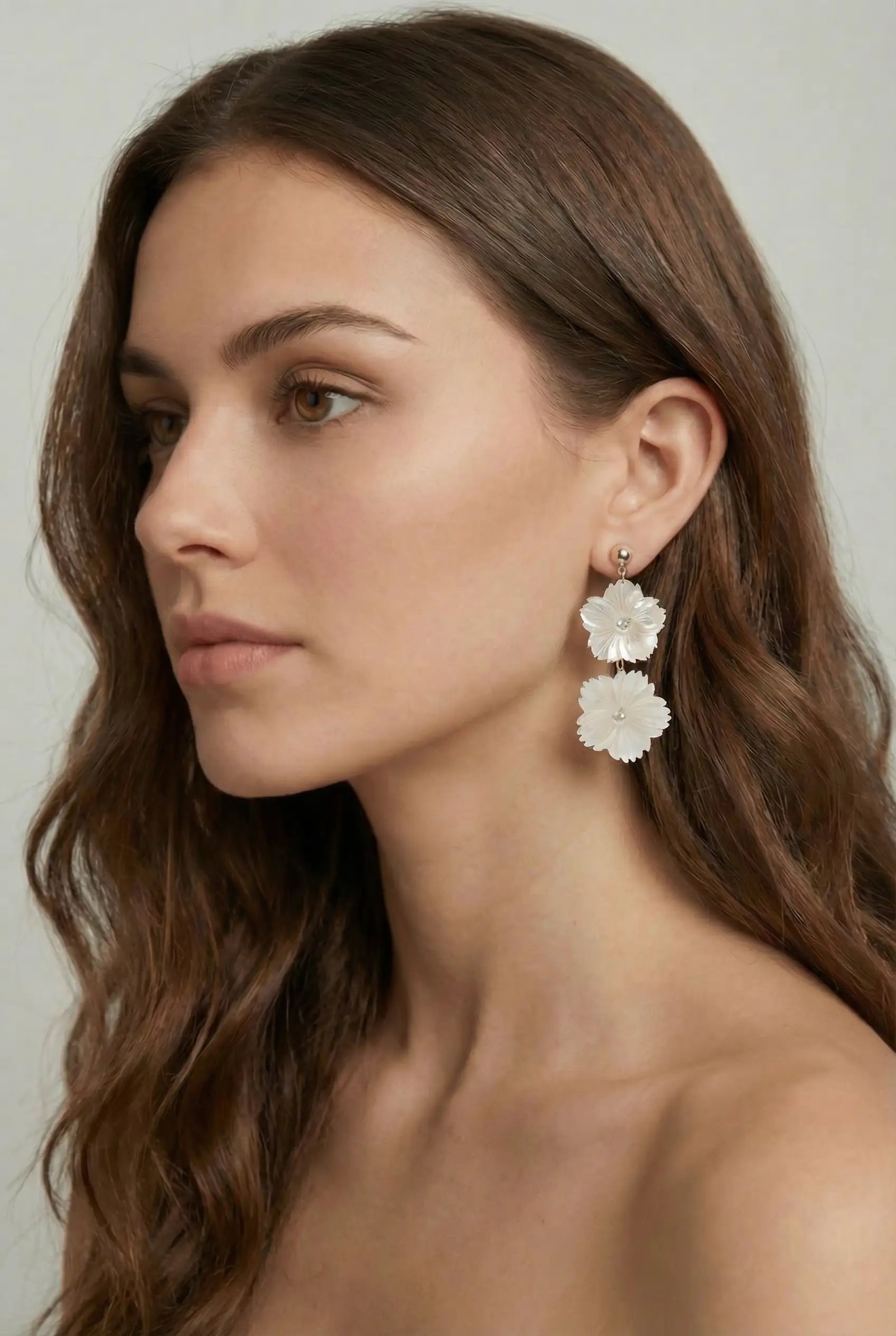 A woman with long, wavy brown hair poses against a neutral background, wearing TH&TH's Pearl Floral Drop Earrings—elegant jewelry evoking timeless bridal style. She gazes slightly left with a calm expression and bare shoulders.