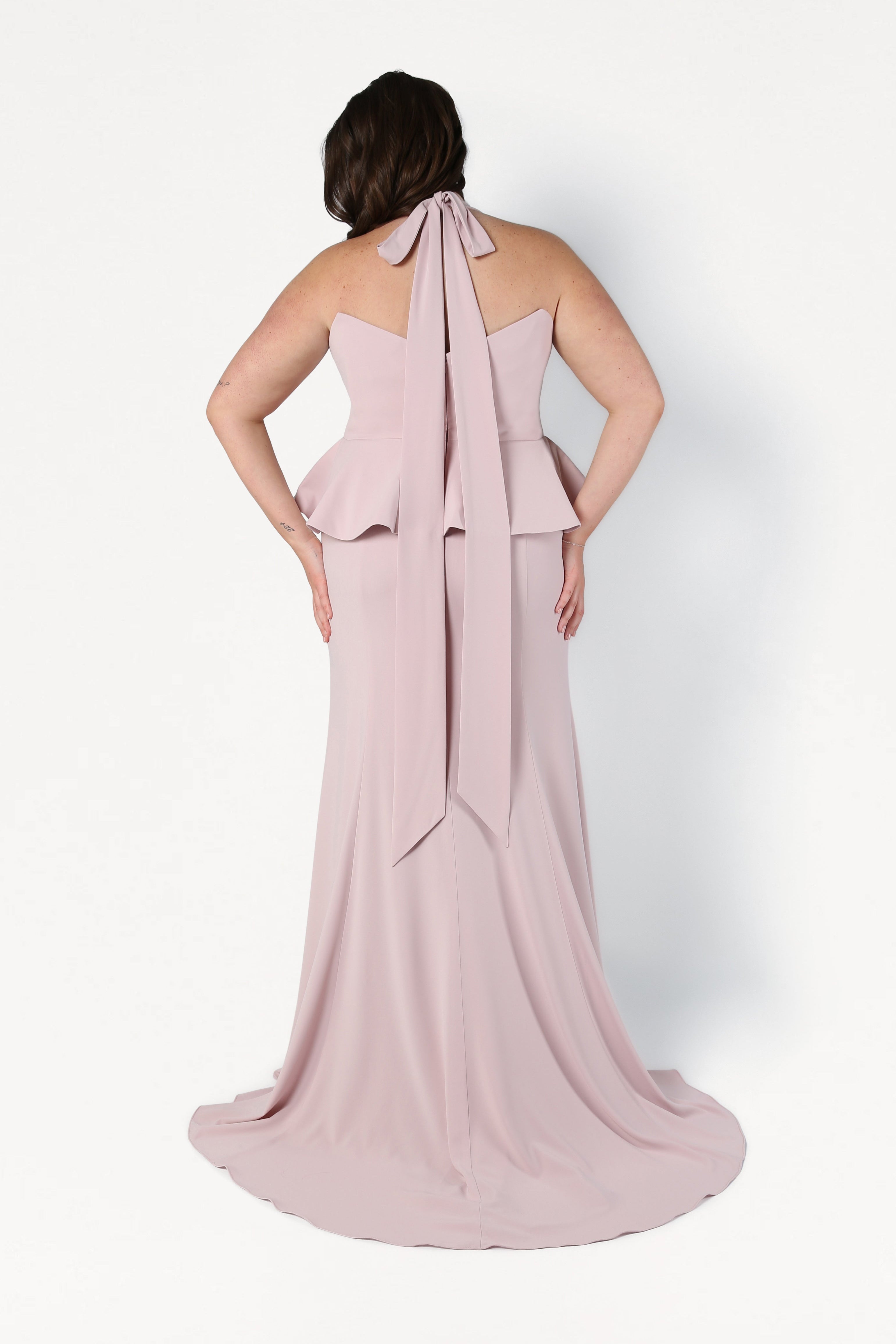 Woman wearing a pink halter neck gown with a large bow detail on a white background