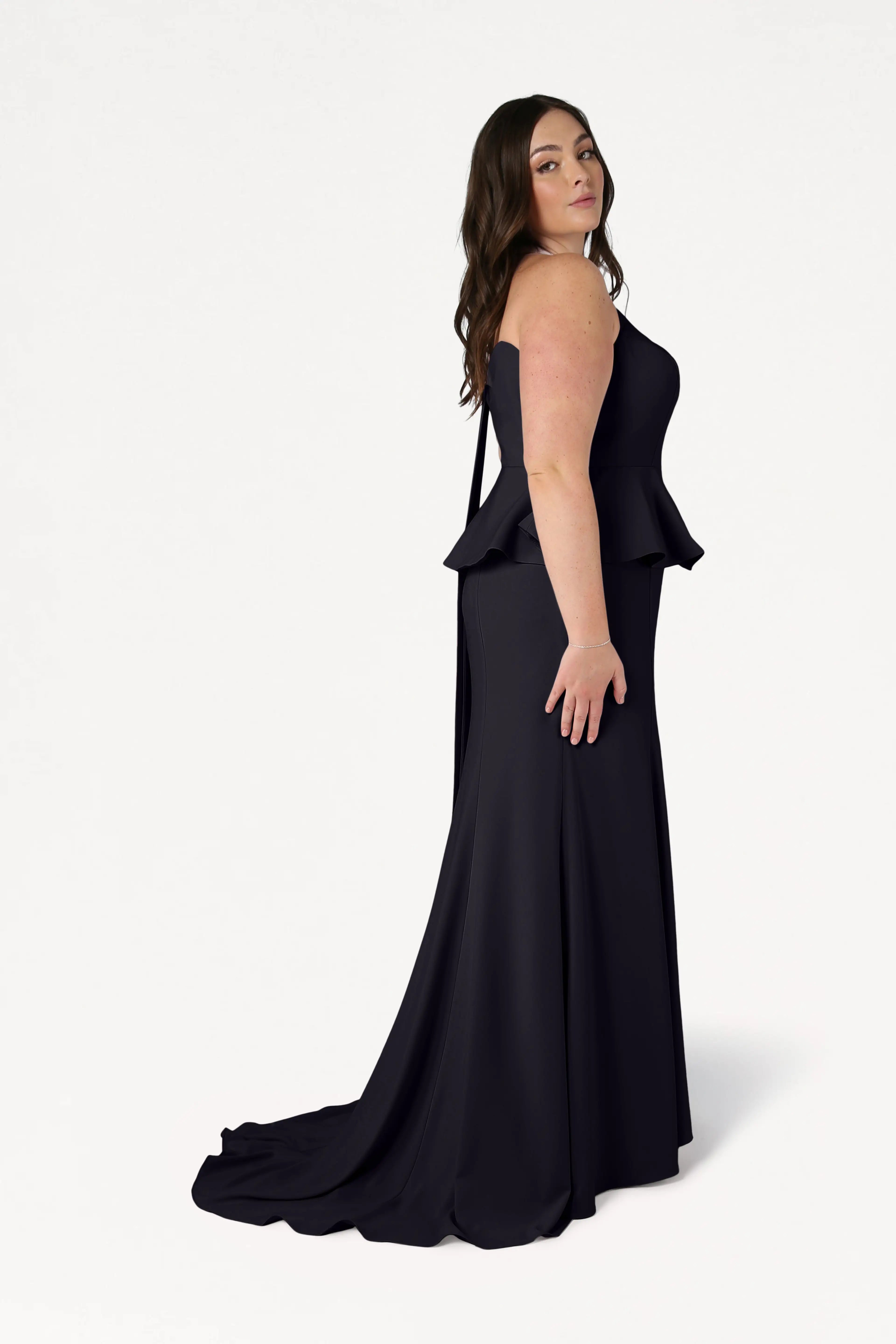 A woman with long brown hair stands in profile against a white background, wearing the TH&TH Peony Bridesmaid Dress in Black—a modern, strapless, floor-length gown featuring peplum detail.