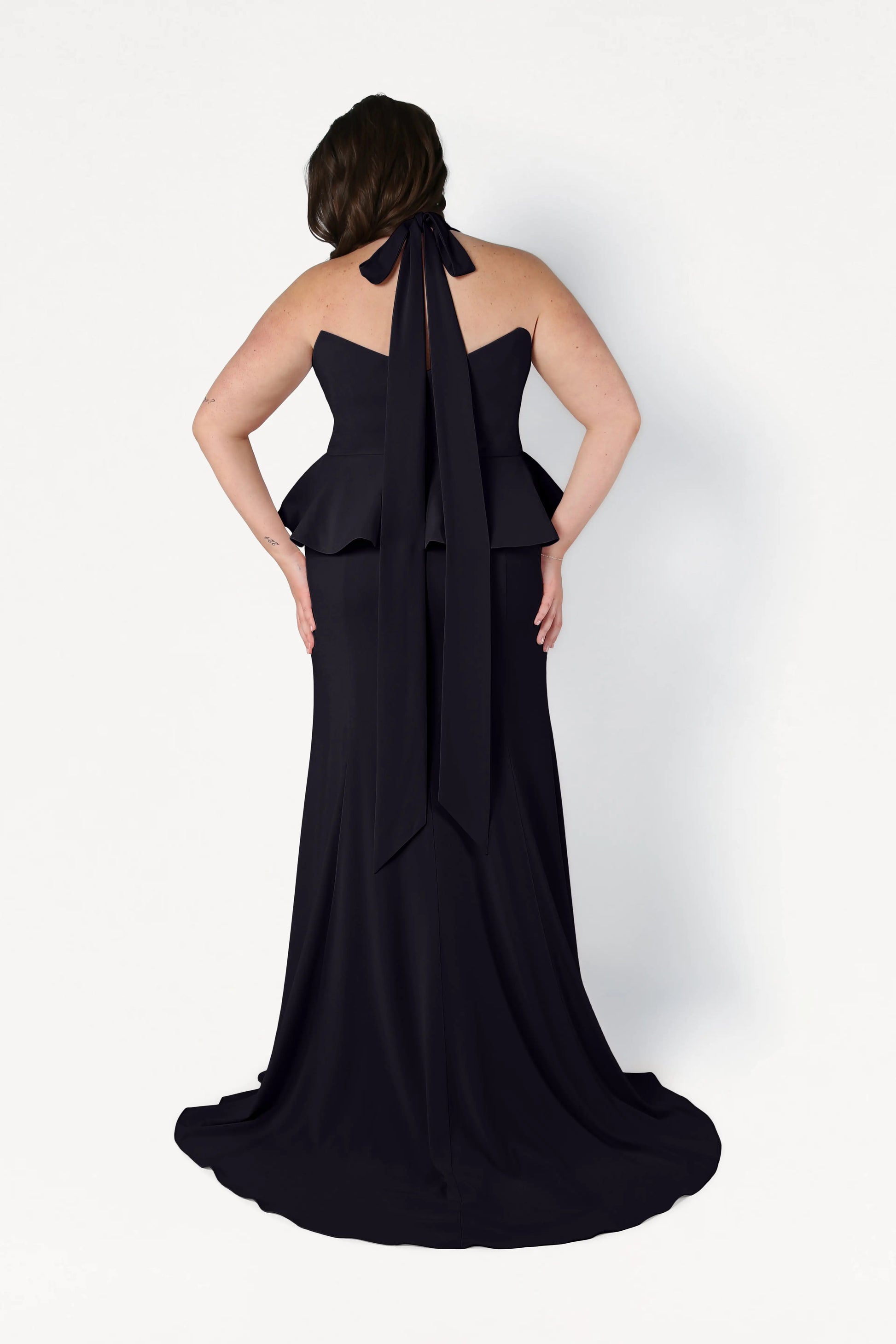 A woman faces away in the TH&TH Peony Bridesmaid Dress in Black—a size-inclusive, modern gown with a peplum waist, halter neck bow, and slight train—standing against a plain white background.