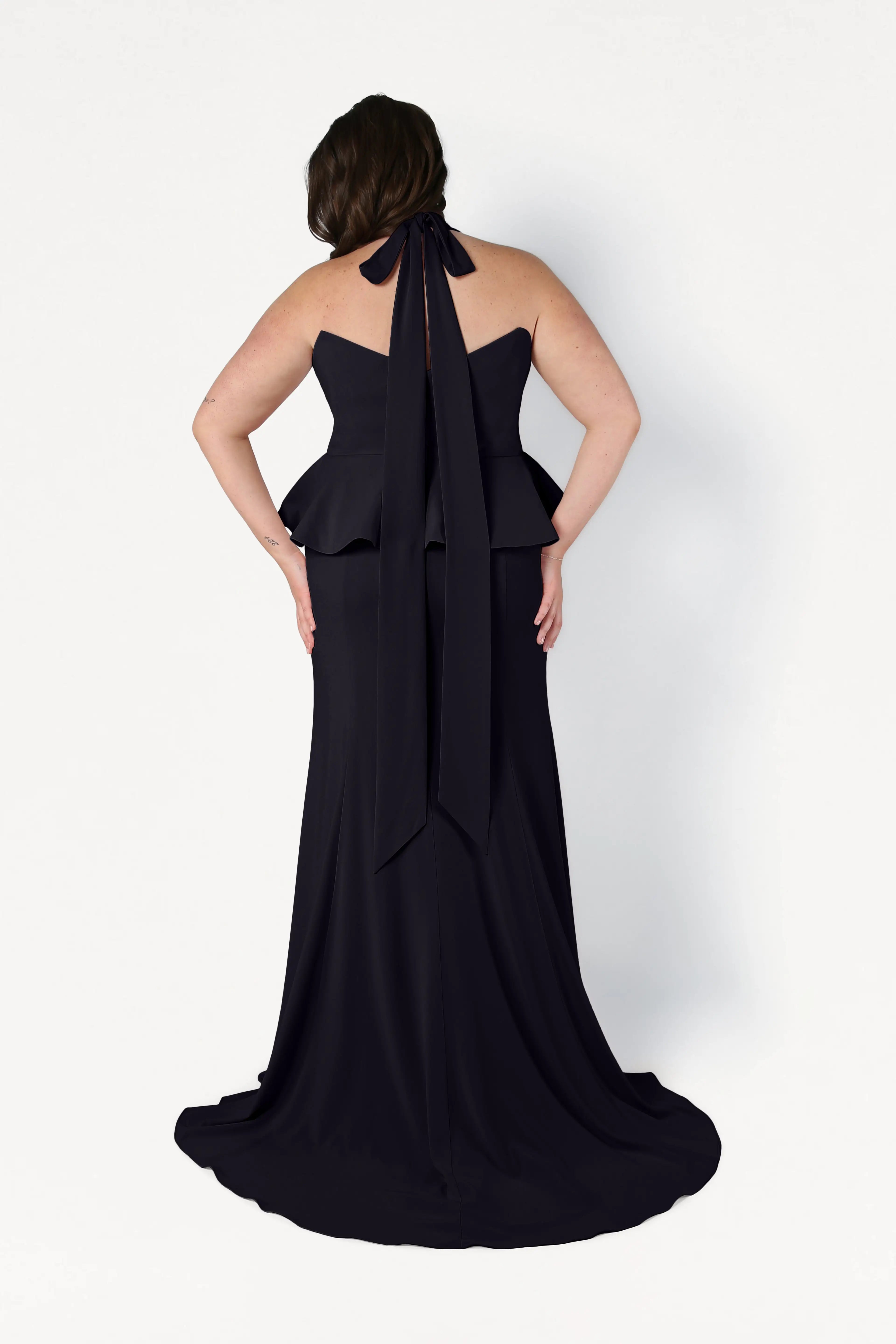 A woman faces away in the TH&TH Peony Bridesmaid Dress in Black—a size-inclusive, modern gown with a peplum waist, halter neck bow, and slight train—standing against a plain white background.
