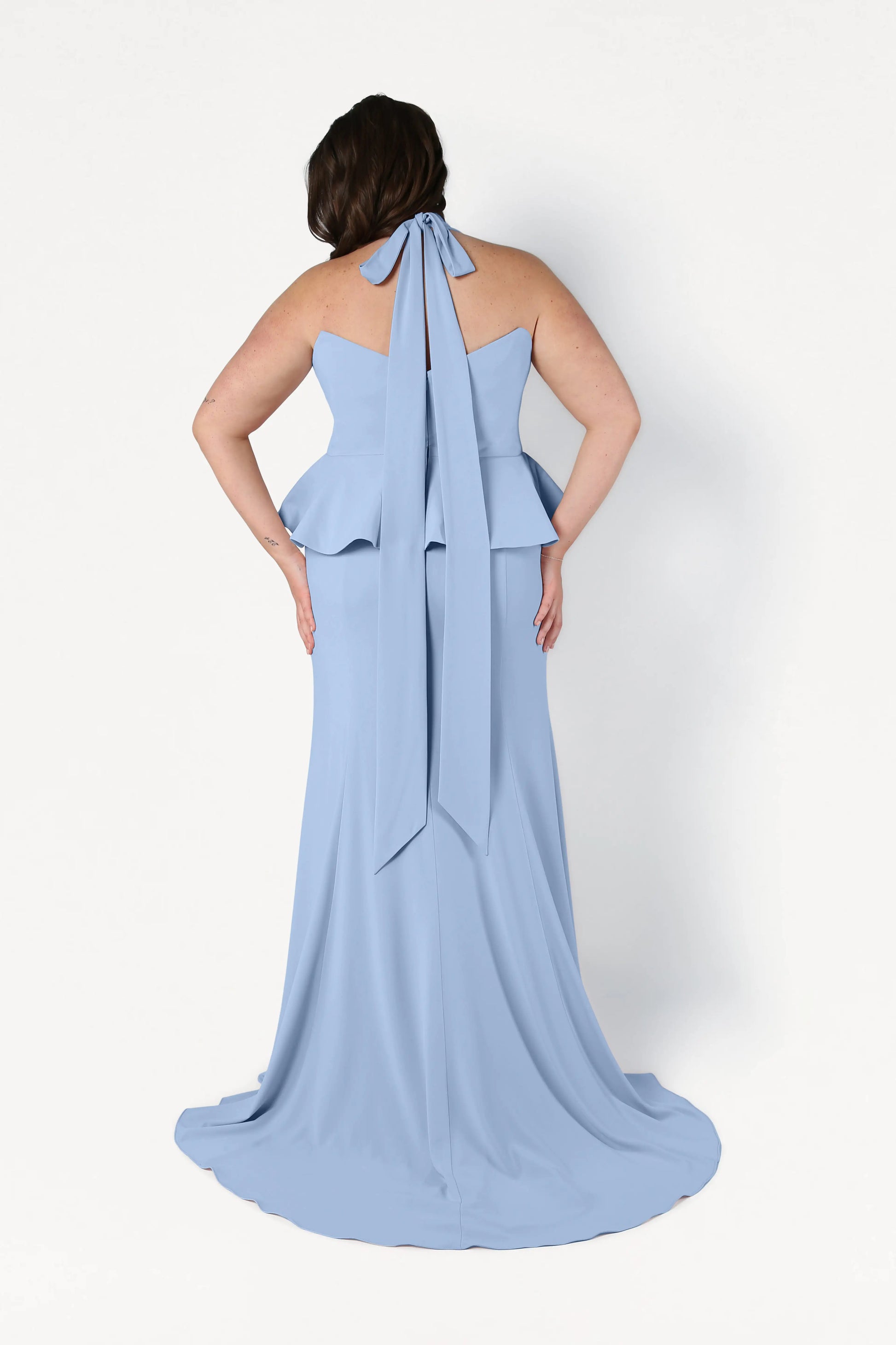 A woman stands facing away in the TH&TH Peony Bridesmaid Dress in Dusty Blue, featuring a peplum waist, back bow tie, and flowing train—a chic, size-inclusive gown against a plain white background.