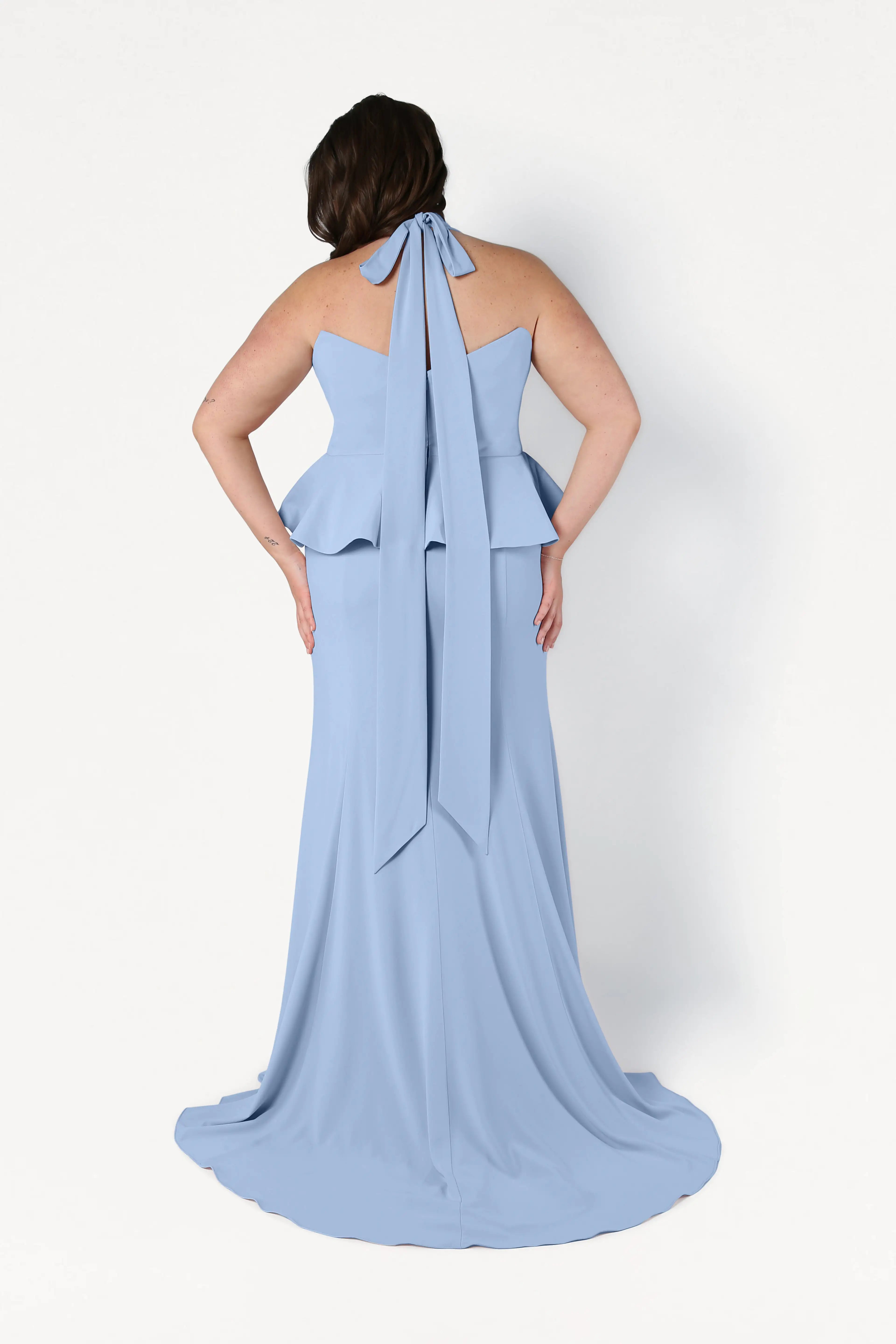 A woman stands facing away in the TH&TH Peony Bridesmaid Dress in Dusty Blue, featuring a peplum waist, back bow tie, and flowing train—a chic, size-inclusive gown against a plain white background.