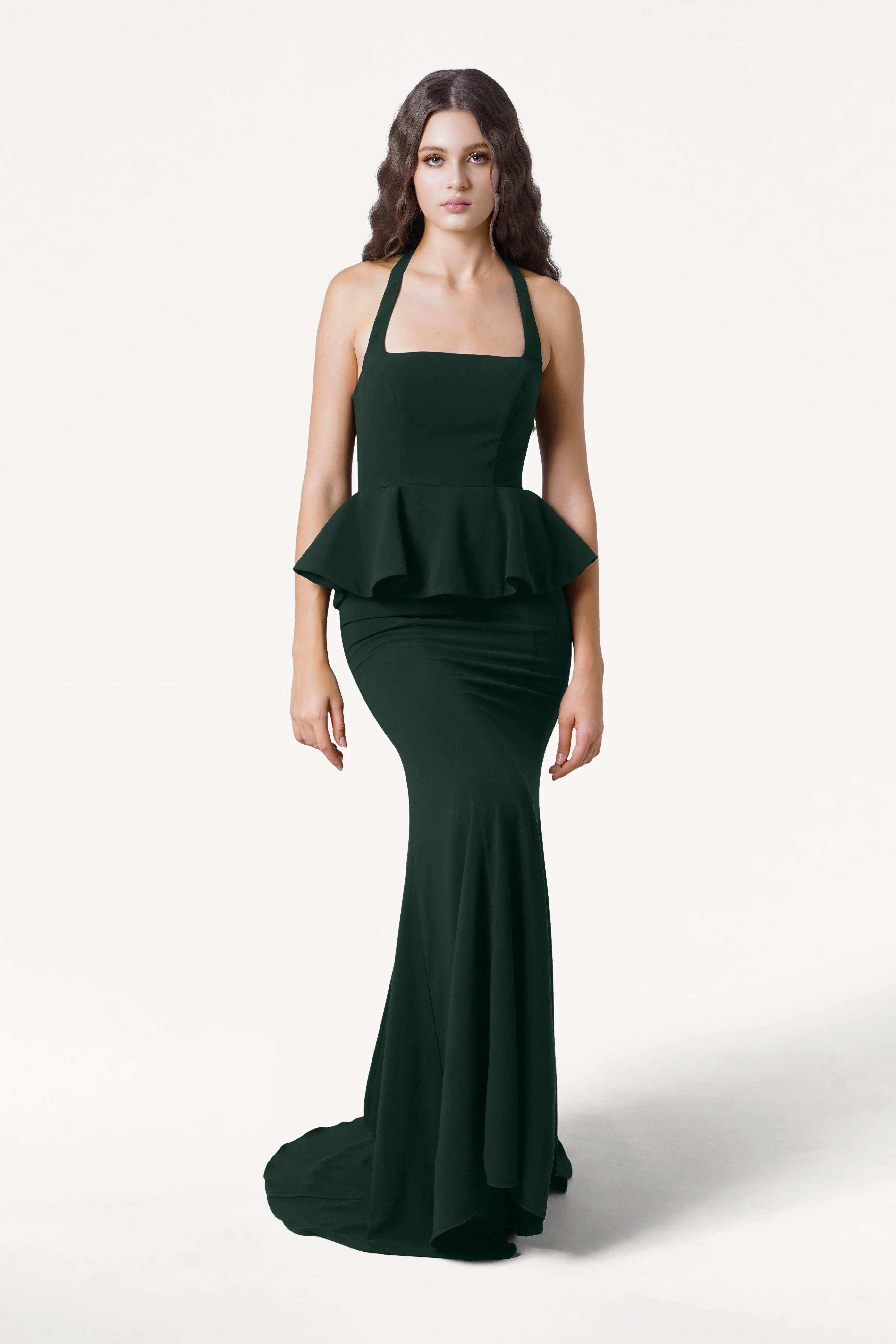 A woman in the TH&TH Peony Bridesmaid Dress in Forest Green, a modern floor-length moss crepe gown with a peplum waist, stands against a white background. Her long, wavy brown hair and neutral expression complete the chic bridal look.