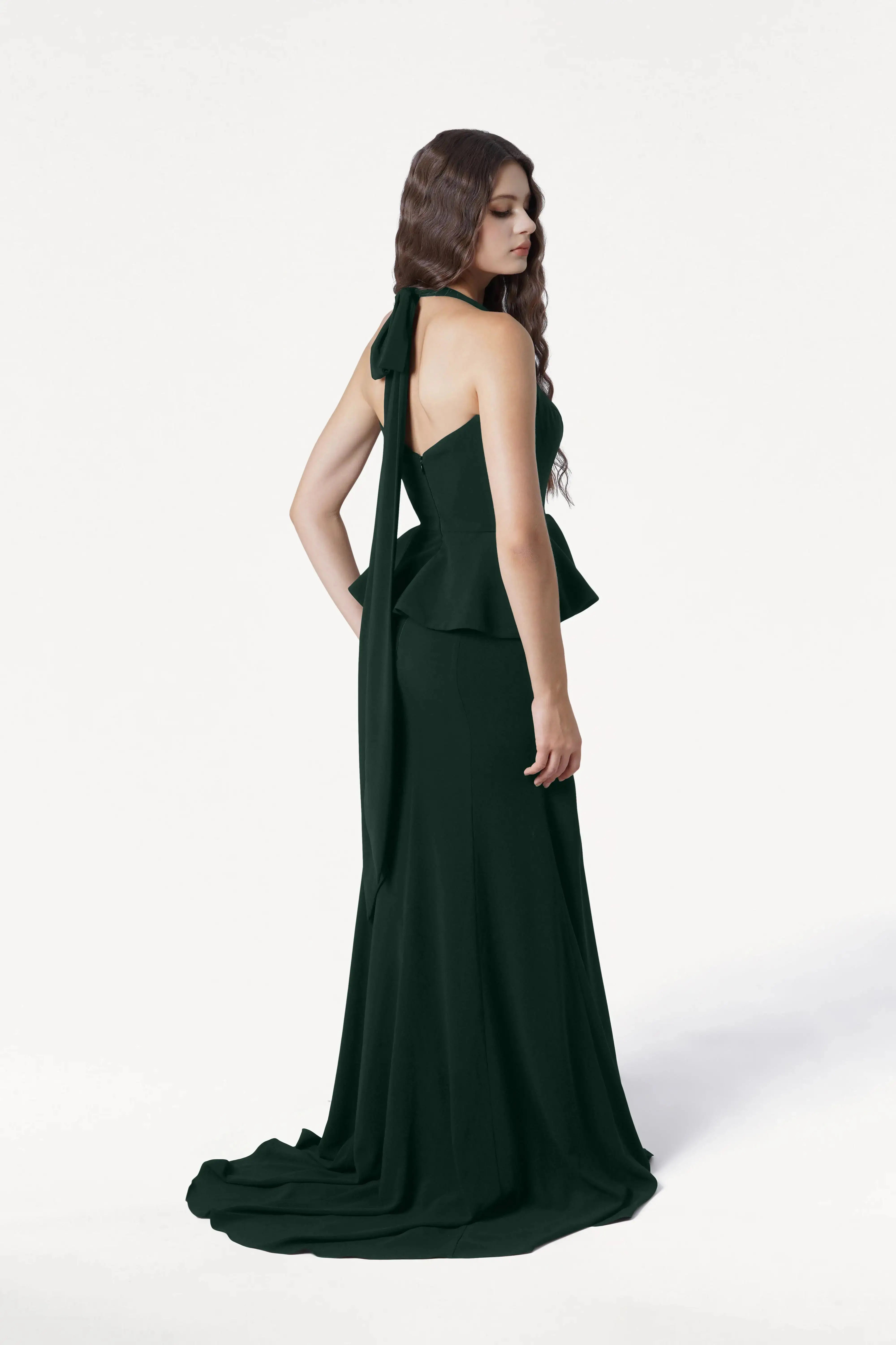 A woman with long wavy brown hair stands in profile, wearing the TH&TH Peony Bridesmaid Dress in Forest Green—a size-inclusive, backless, floor-length crepe gown with a flowing train and large neck bow—perfect for modern weddings.
