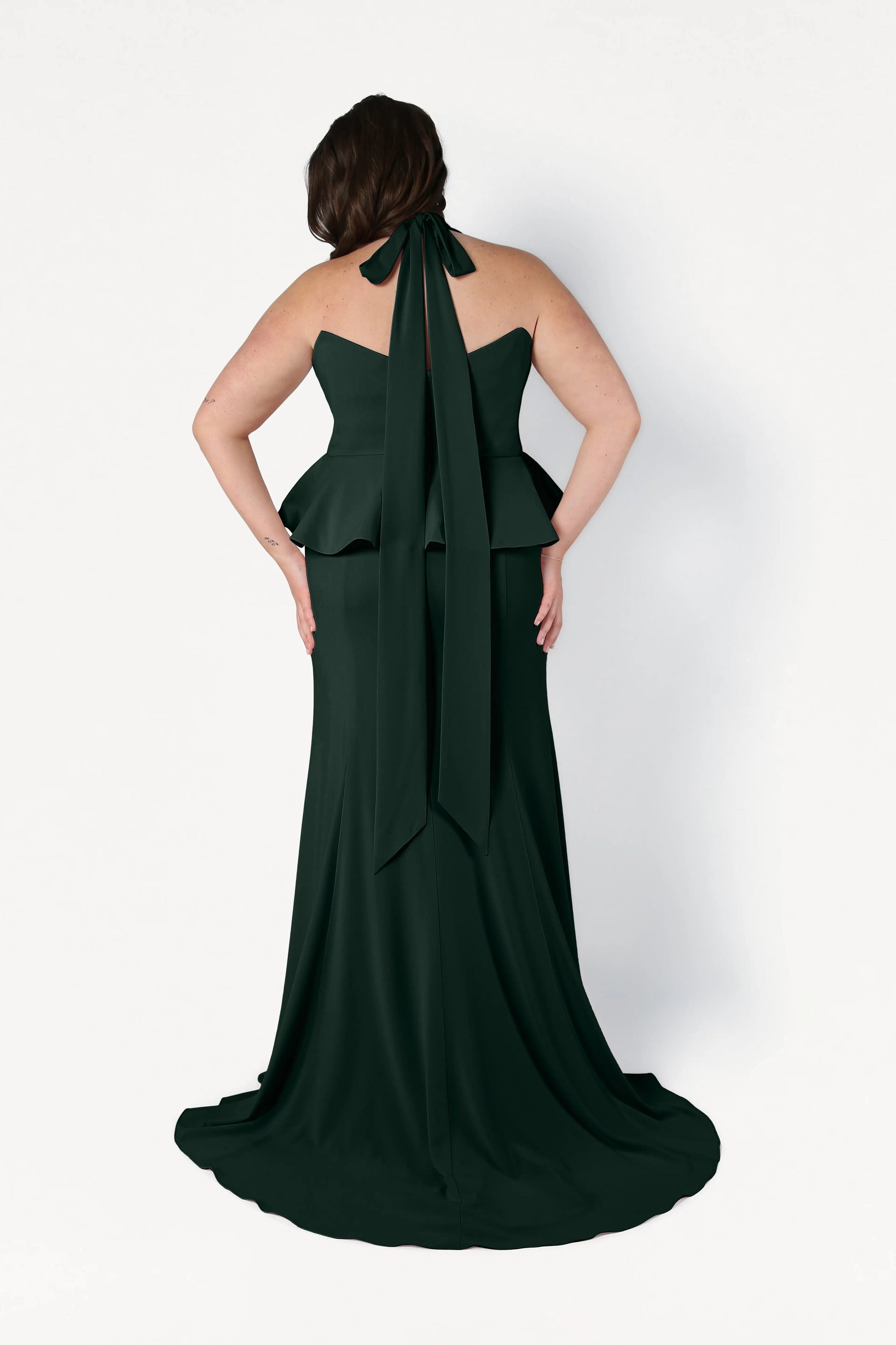 A woman wears the size-inclusive TH&TH Peony Bridesmaid Dress in Forest Green crepe, featuring a peplum waist and statement neck bow, shown from the back against a white background. Ideal for contemporary weddings.