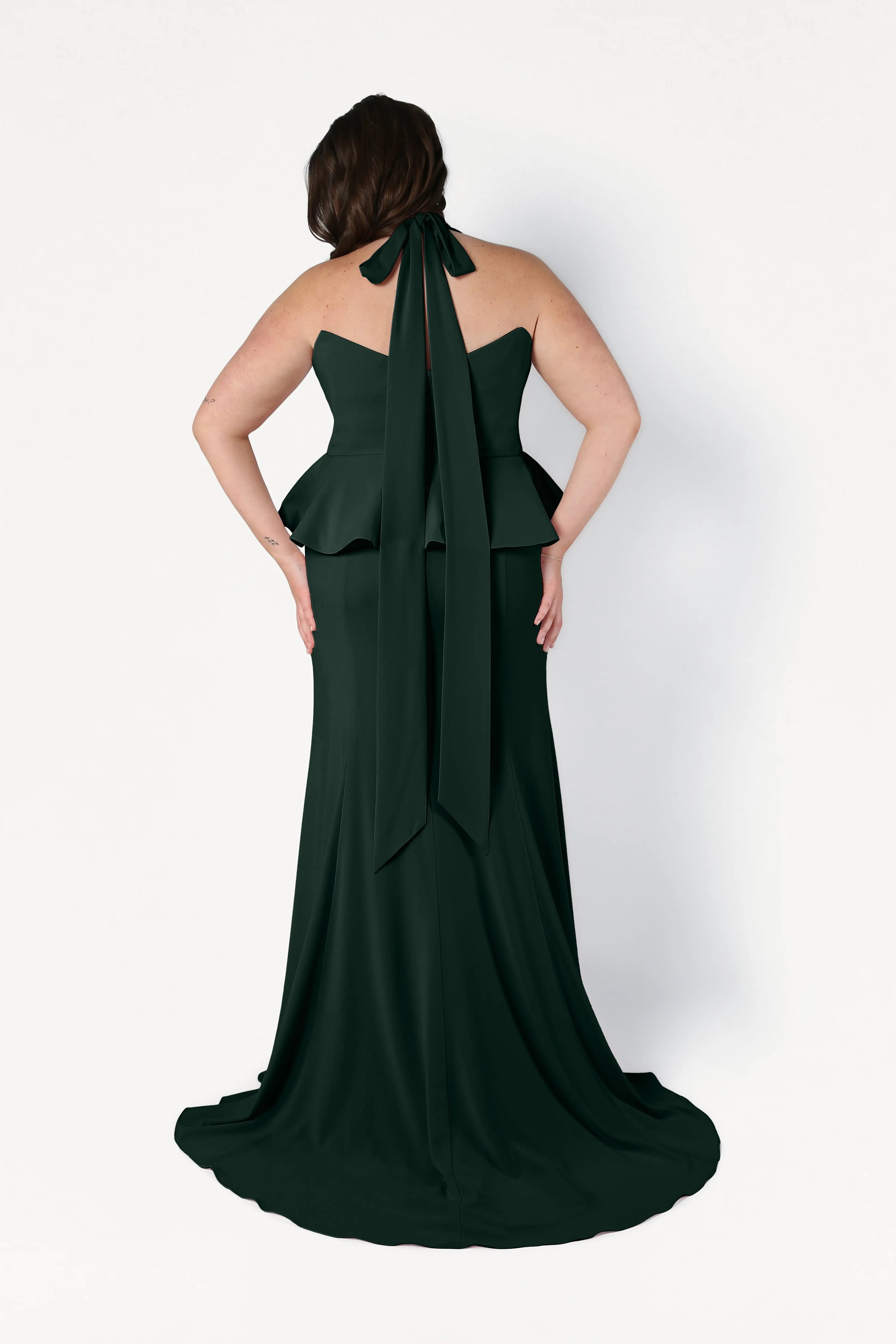 A woman wears the size-inclusive TH&TH Peony Bridesmaid Dress in Forest Green crepe, featuring a peplum waist and statement neck bow, shown from the back against a white background. Ideal for contemporary weddings.