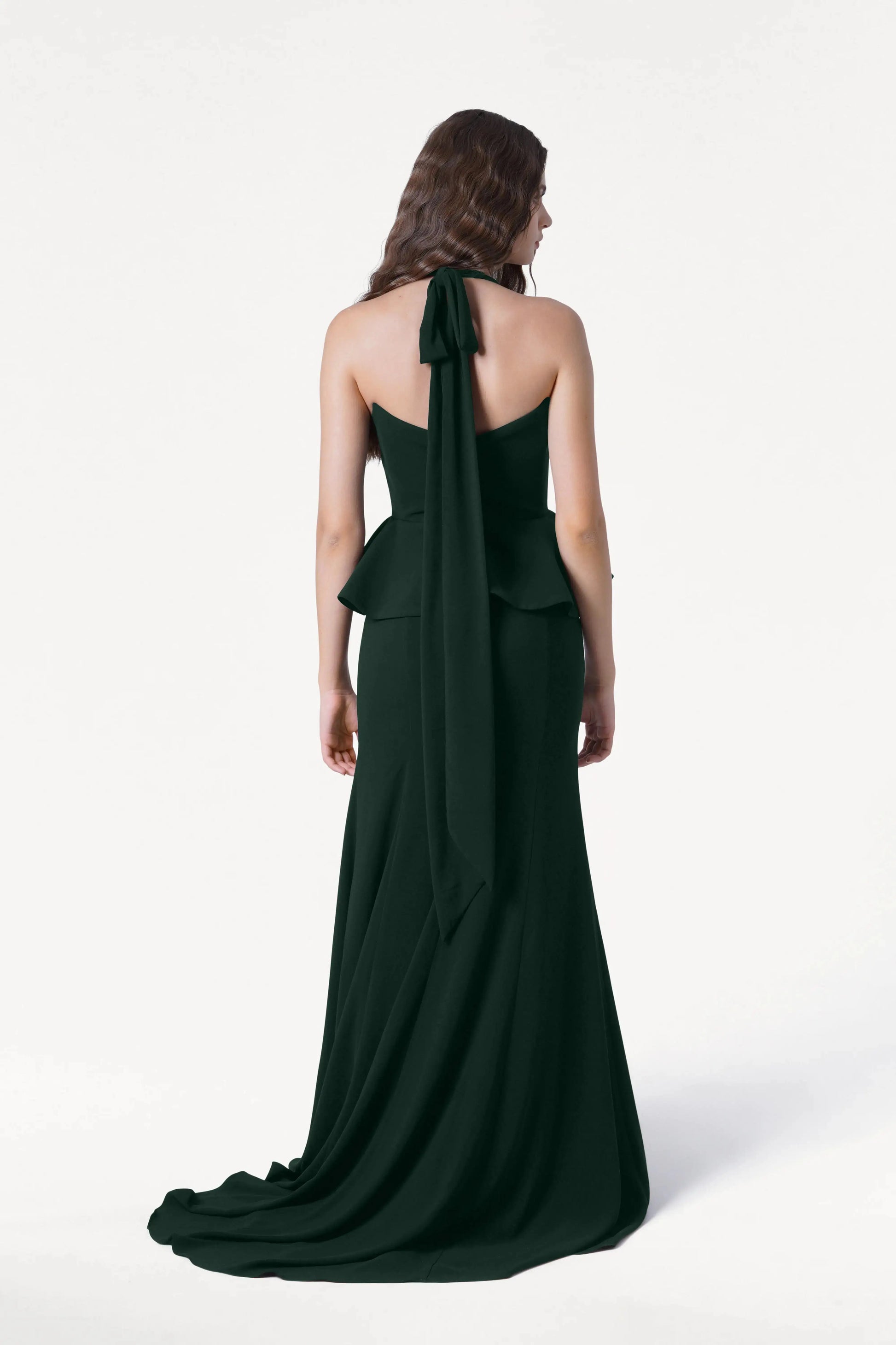 A woman with wavy brown hair stands facing away, wearing the TH&TH Peony Bridesmaid Dress in Forest Green with a peplum waist and long halter tie, ideal for modern weddings, against a plain white background.