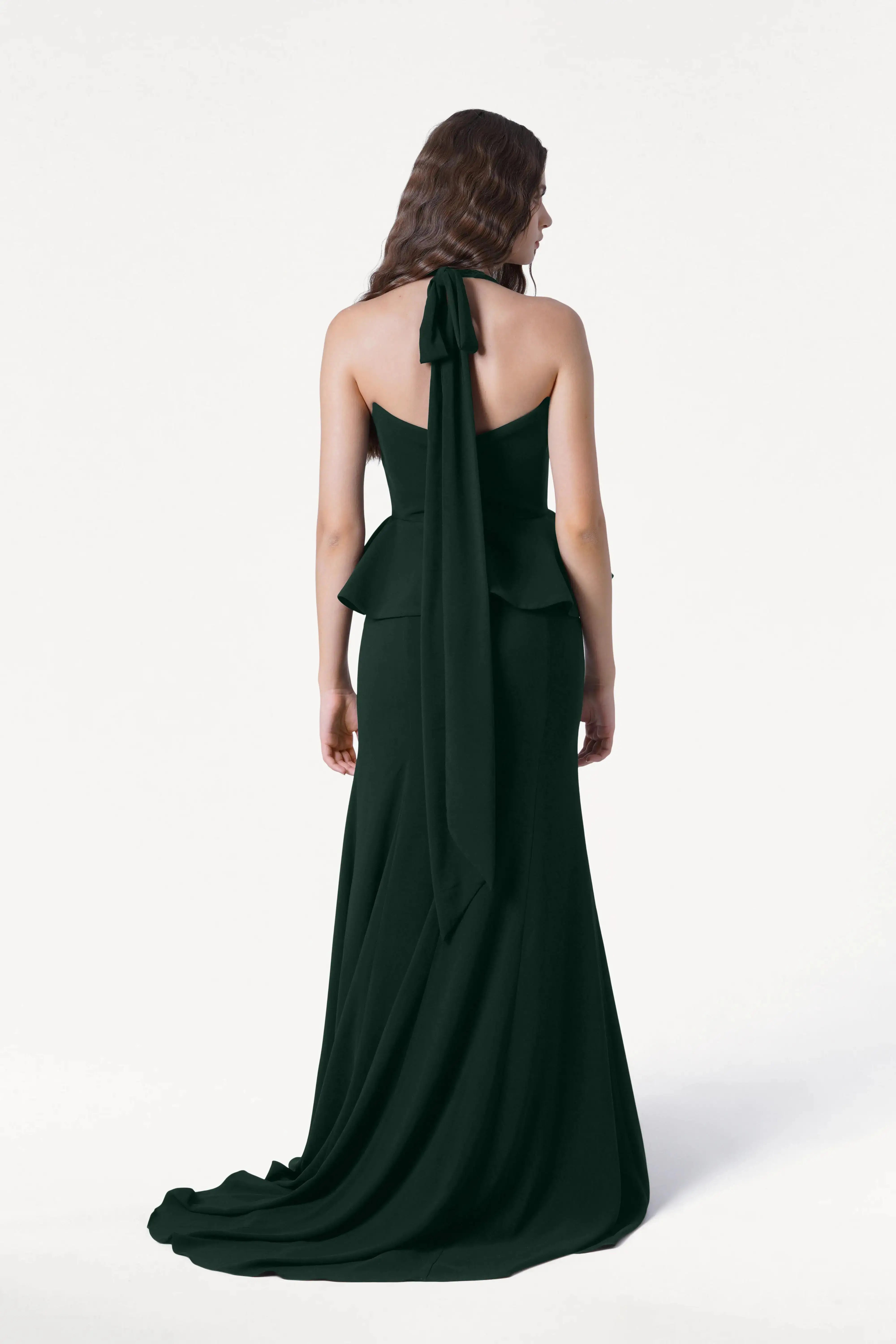A woman with wavy brown hair stands facing away, wearing the TH&TH Peony Bridesmaid Dress in Forest Green with a peplum waist and long halter tie, ideal for modern weddings, against a plain white background.