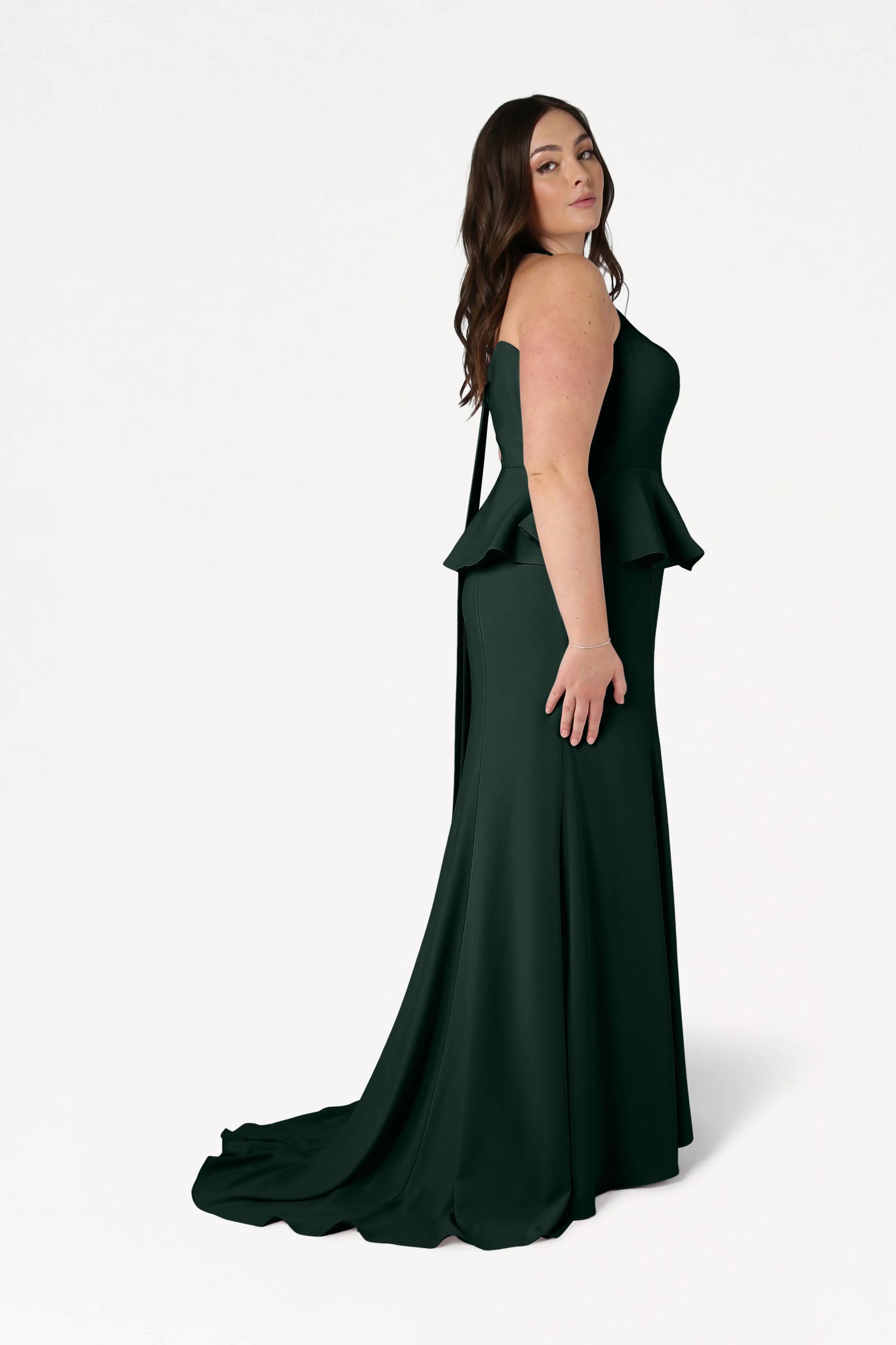 A woman stands sideways against a white background in the Peony Bridesmaid Dress in Forest Green by TH&TH—a strapless, floor-length modern gown with peplum detail—looking confidently at the camera, her loose waves perfect for contemporary weddings.