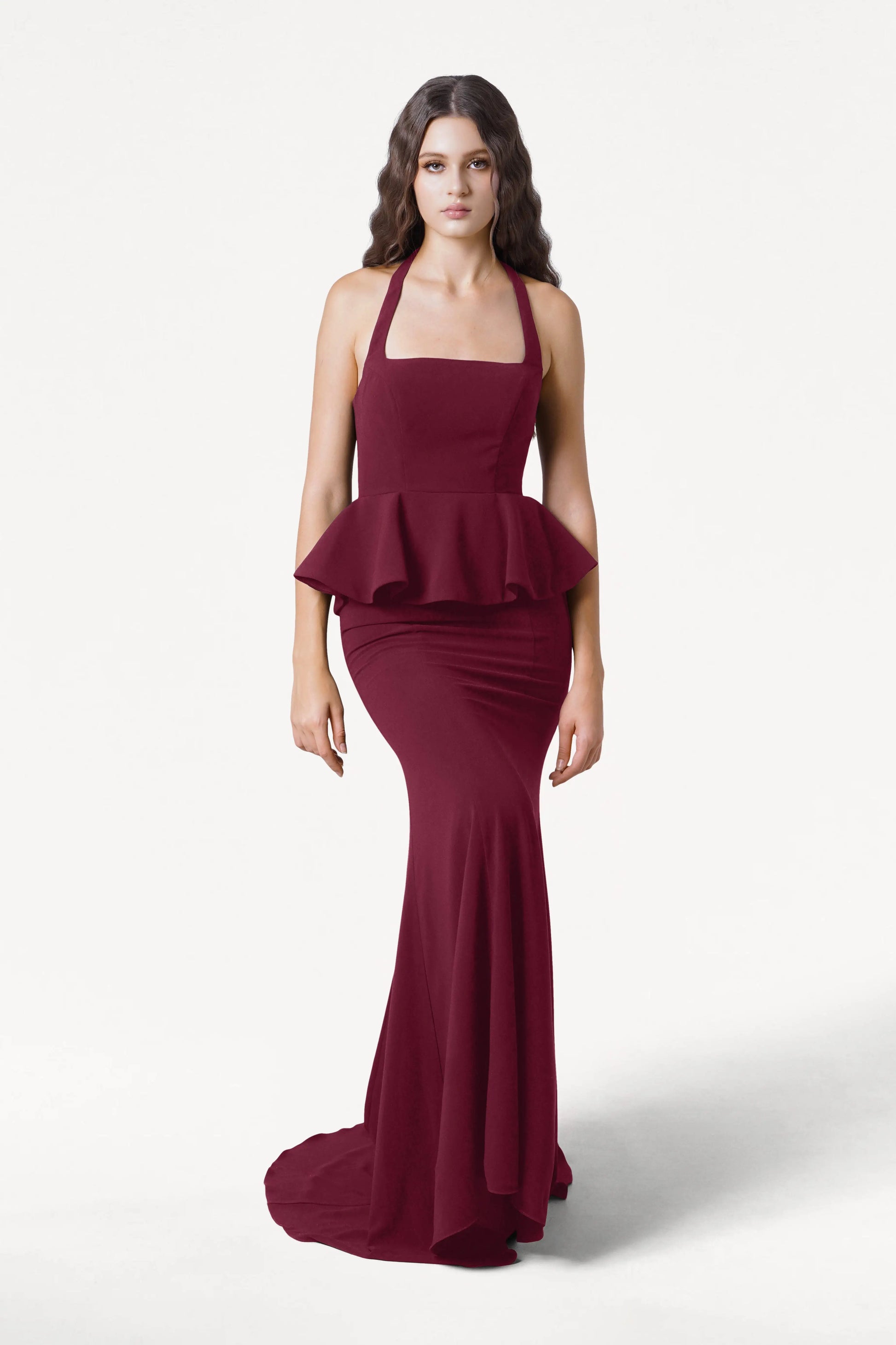 A woman with long, wavy dark hair and a neutral expression stands against a plain white background, modeling the TH&TH Peony Bridesmaid Dress in Merlot—a sleeveless, floor-length gown with a peplum waist.