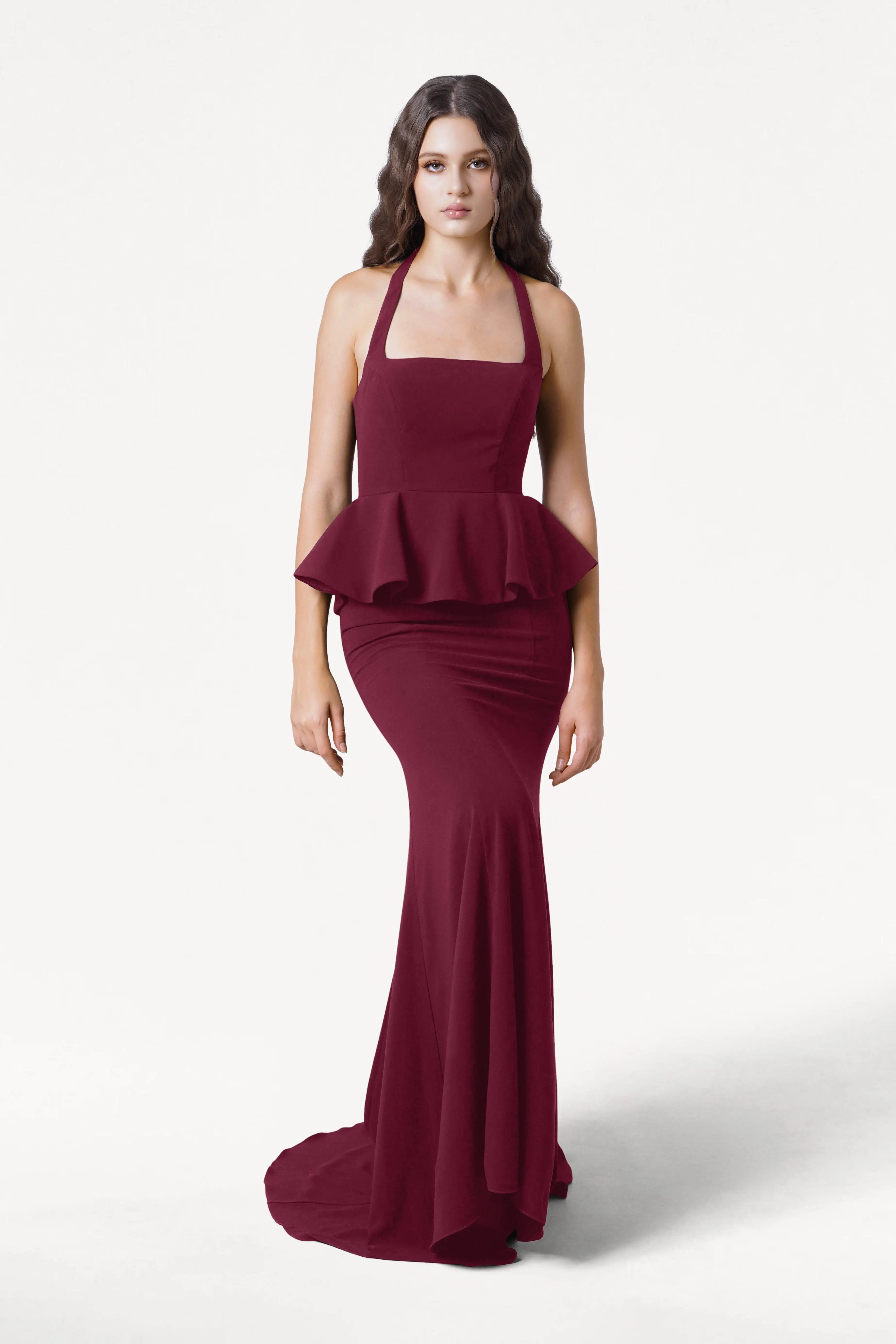 A woman with long, wavy dark hair and a neutral expression stands against a plain white background, modeling the TH&TH Peony Bridesmaid Dress in Merlot—a sleeveless, floor-length gown with a peplum waist.