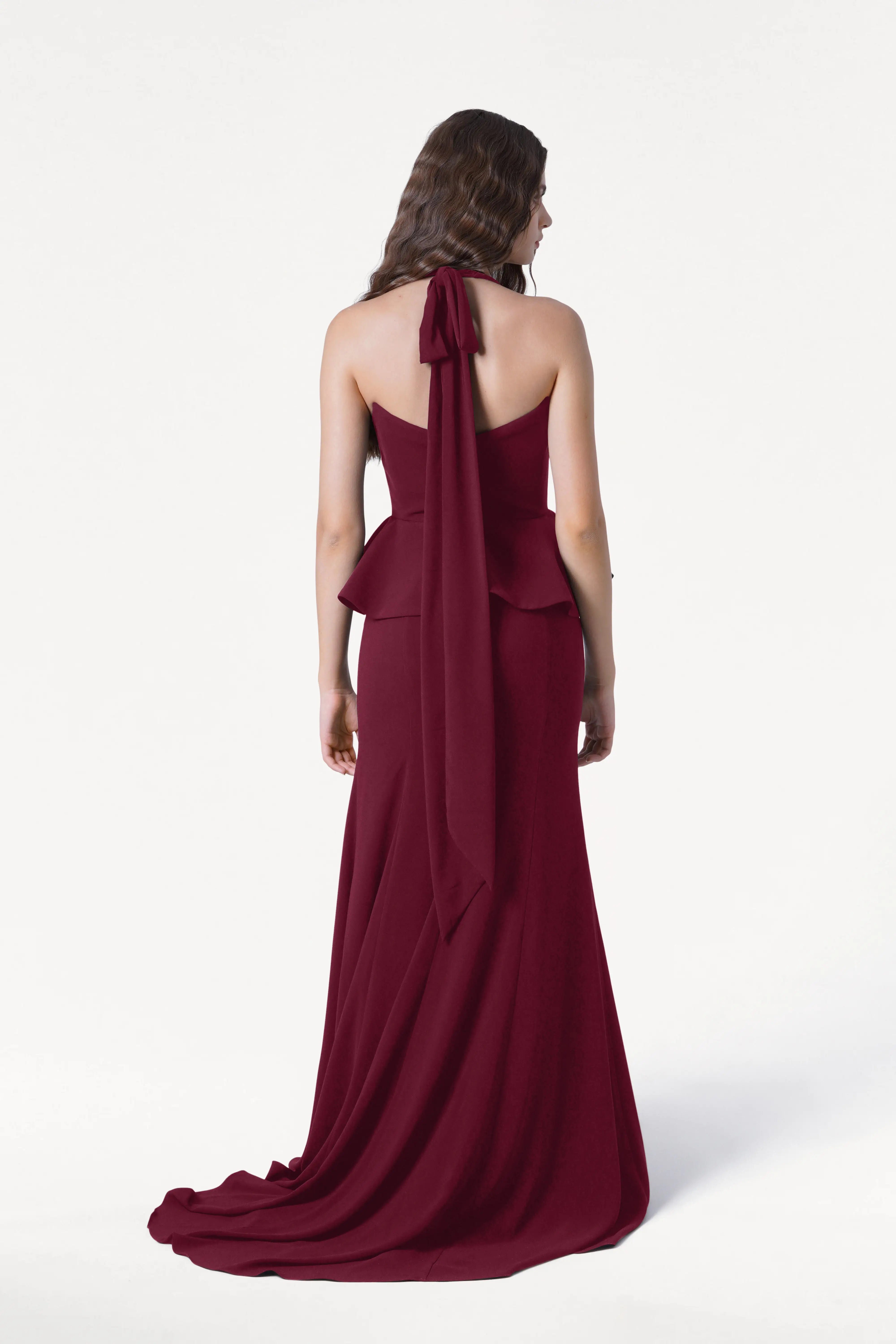 A woman with long brown hair stands with her back to the camera against a white background, wearing the TH&TH Peony Bridesmaid Dress in Merlot, a floor-length gown with a sash and peplum detail.