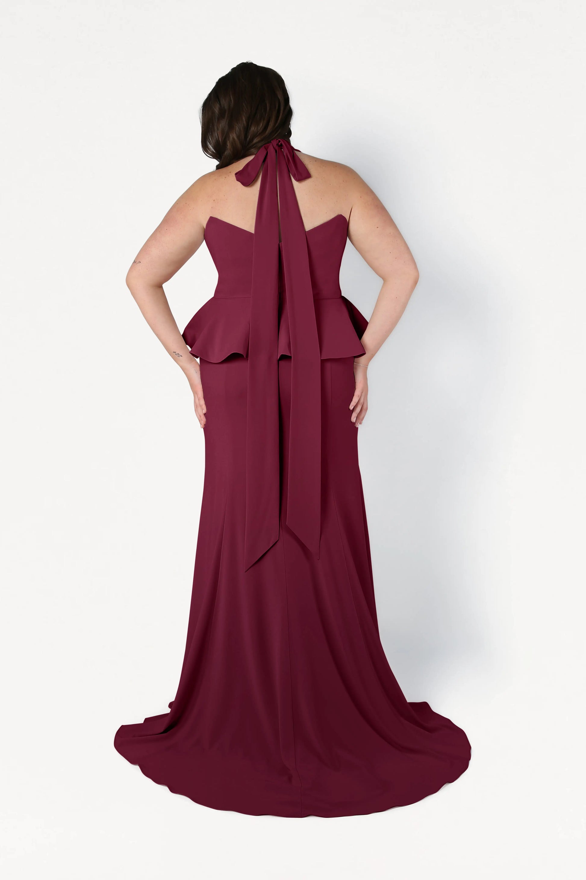 A woman faces away in a TH&TH Peony Bridesmaid Dress in Merlot, featuring a floor-length, sleeveless design with a peplum waist and a long neck bow, set against a plain white background.