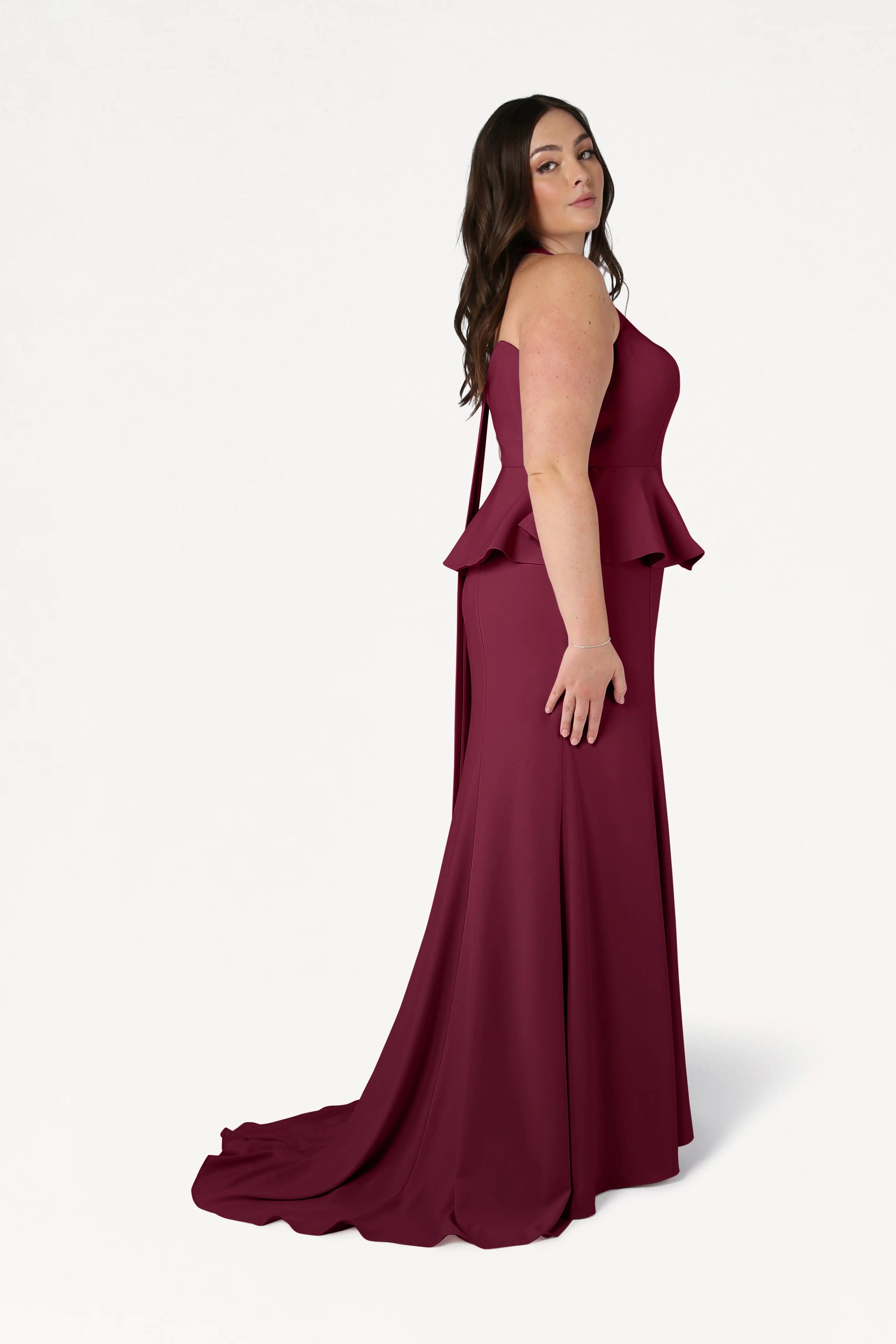 A woman with long, wavy dark hair stands sideways against a white background, confidently modeling the TH&TH Peony Bridesmaid Dress in Merlot—a sleeveless, floor-length gown with a peplum waist and slight train.