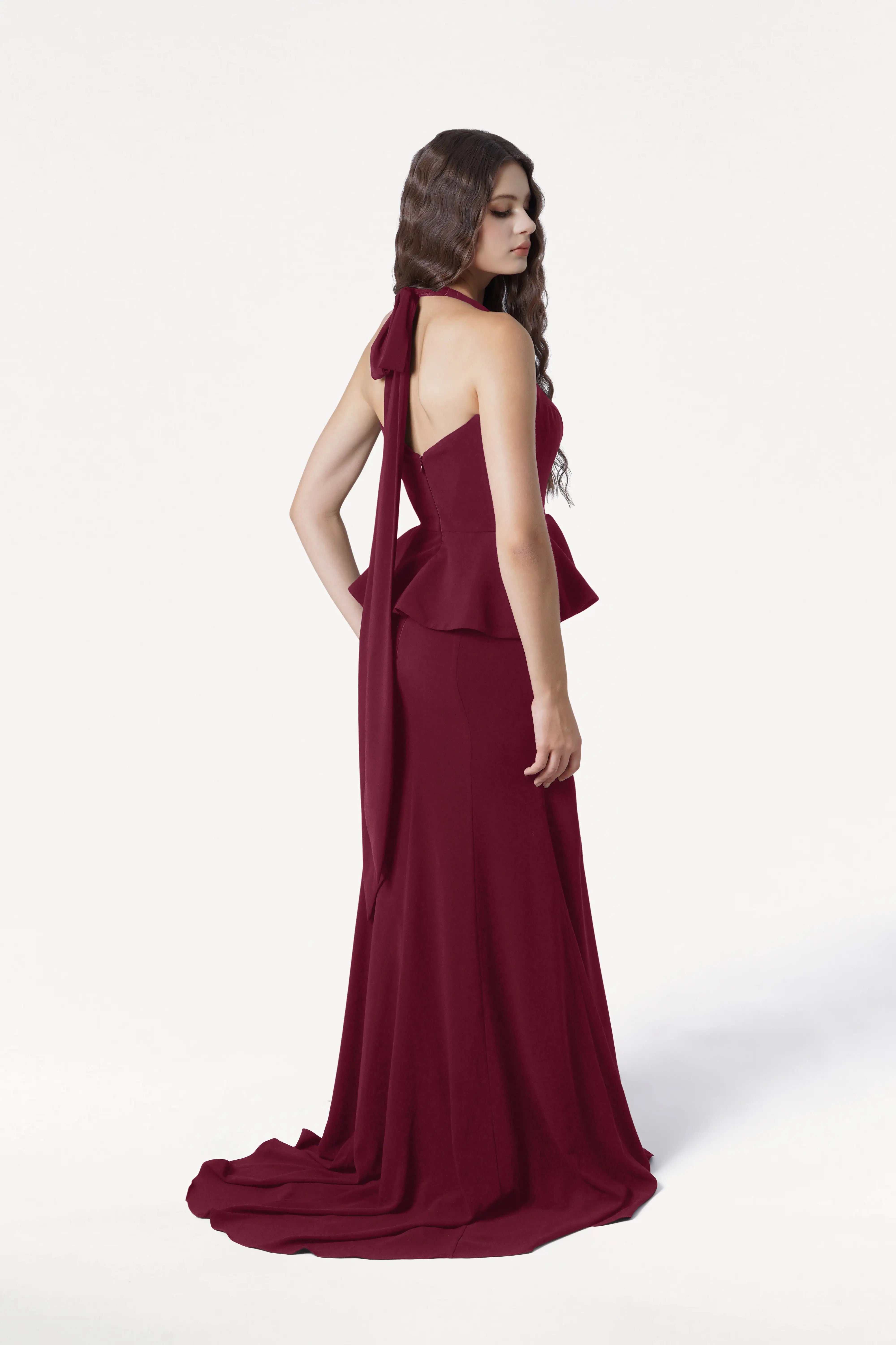 A woman stands in a side pose against a plain white background, wearing the Peony Bridesmaid Dress in Merlot by TH&TH, featuring elegant moss crepe fabric, a peplum waist, and a bow-tied halter neckline.