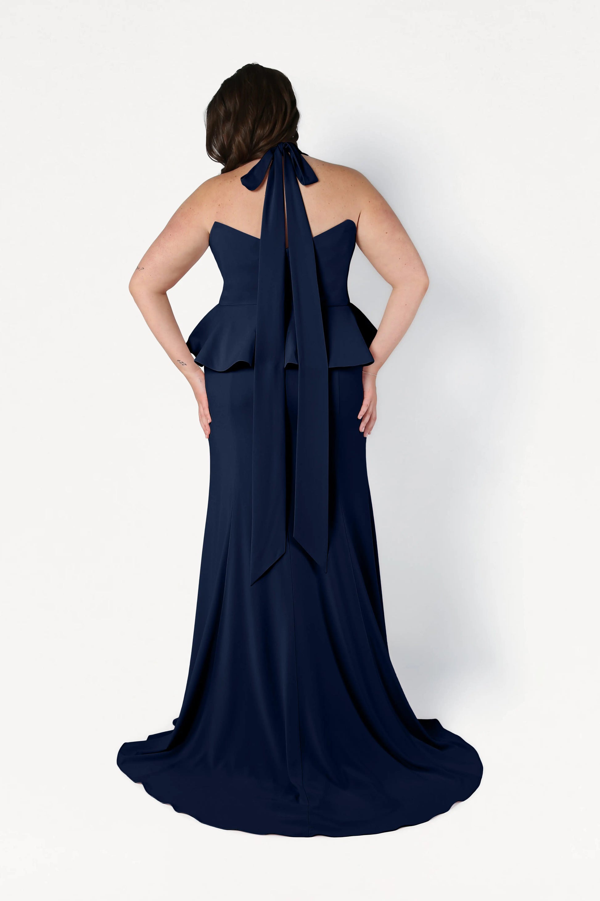 A woman is shown from the back wearing the Peony Bridesmaid Dress in Navy by TH&TH, featuring a floor-length design with a peplum waist and halter neck bow, set against a plain white background.