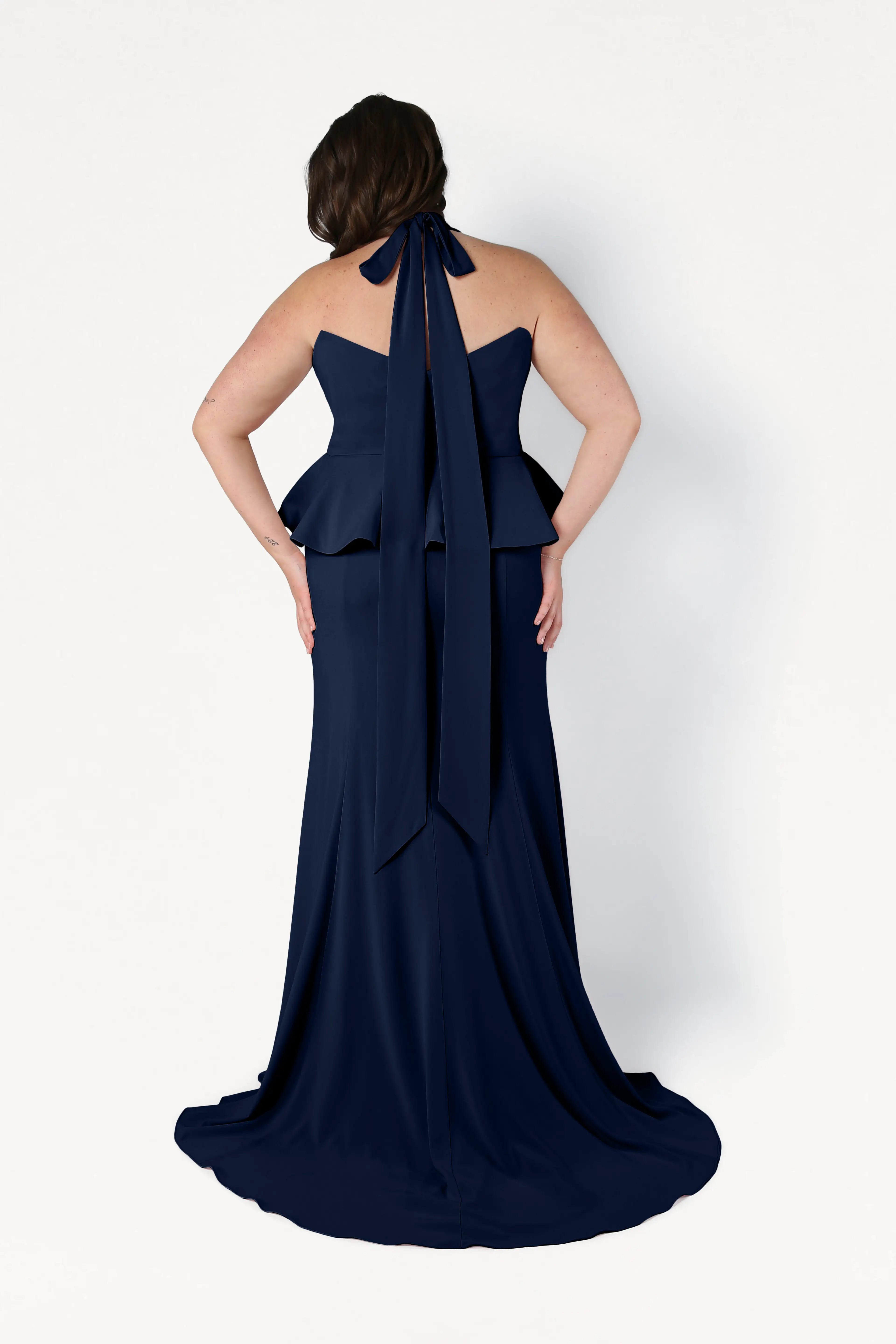 A woman is shown from the back wearing the Peony Bridesmaid Dress in Navy by TH&TH, featuring a floor-length design with a peplum waist and halter neck bow, set against a plain white background.